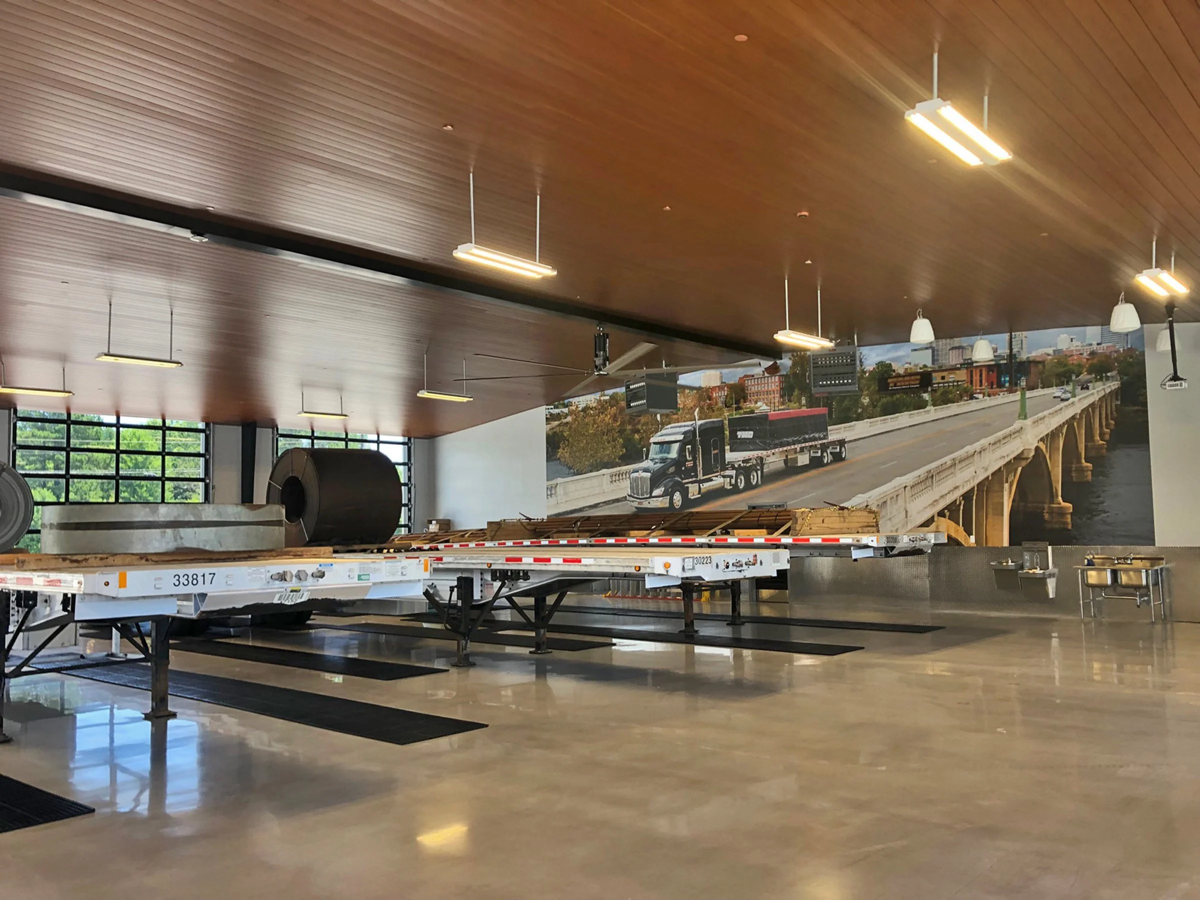 Interior view of a modern building with a wooden ceiling, hanging lights, a large window, and a mural or photo of a city scene including a bridge and trucks outside.