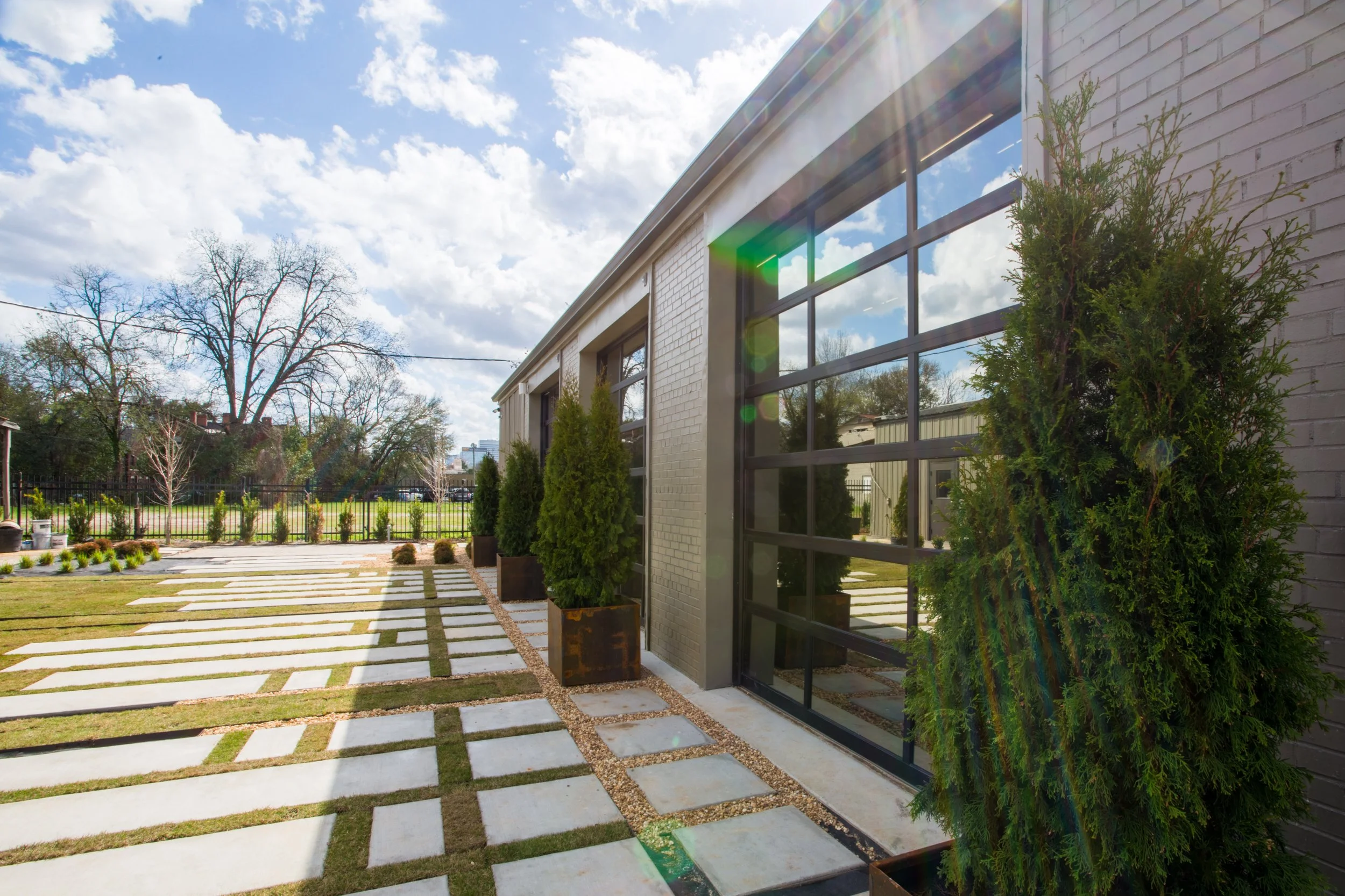Modern building with large glass windows, surrounded by small potted evergreen trees, with a decorative pathway and a fenced yard, under a partly cloudy sky with sunlight lens flare.