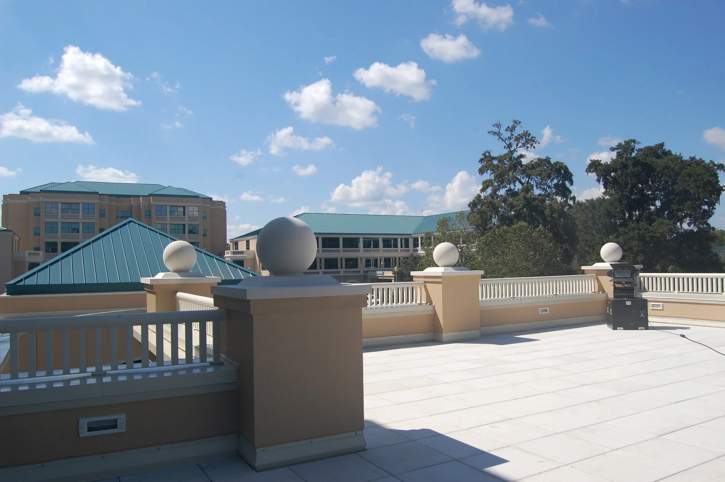 View of a rooftop terrace with a beige railing and decorative white spheres, with a building with a blue roof and trees in the background, under a partly cloudy sky.