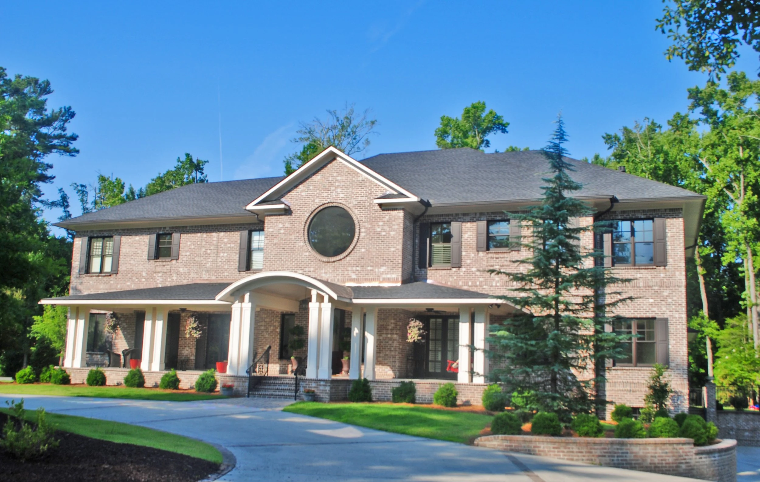Large brick house with a circular window above the porch, surrounded by green trees and a well-maintained lawn, under a clear blue sky.