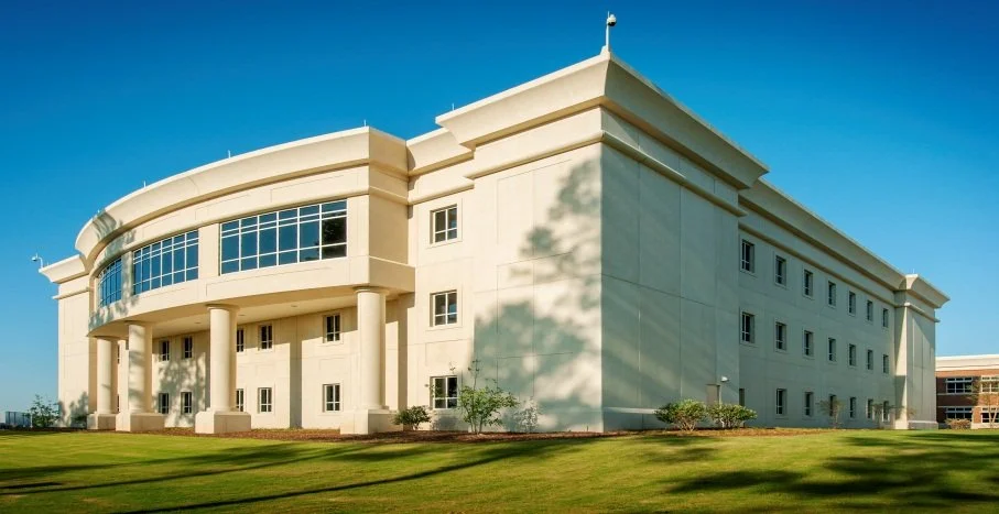Large modern beige building with curved glass windows and tall columns, surrounded by green grass and trees under a clear blue sky.