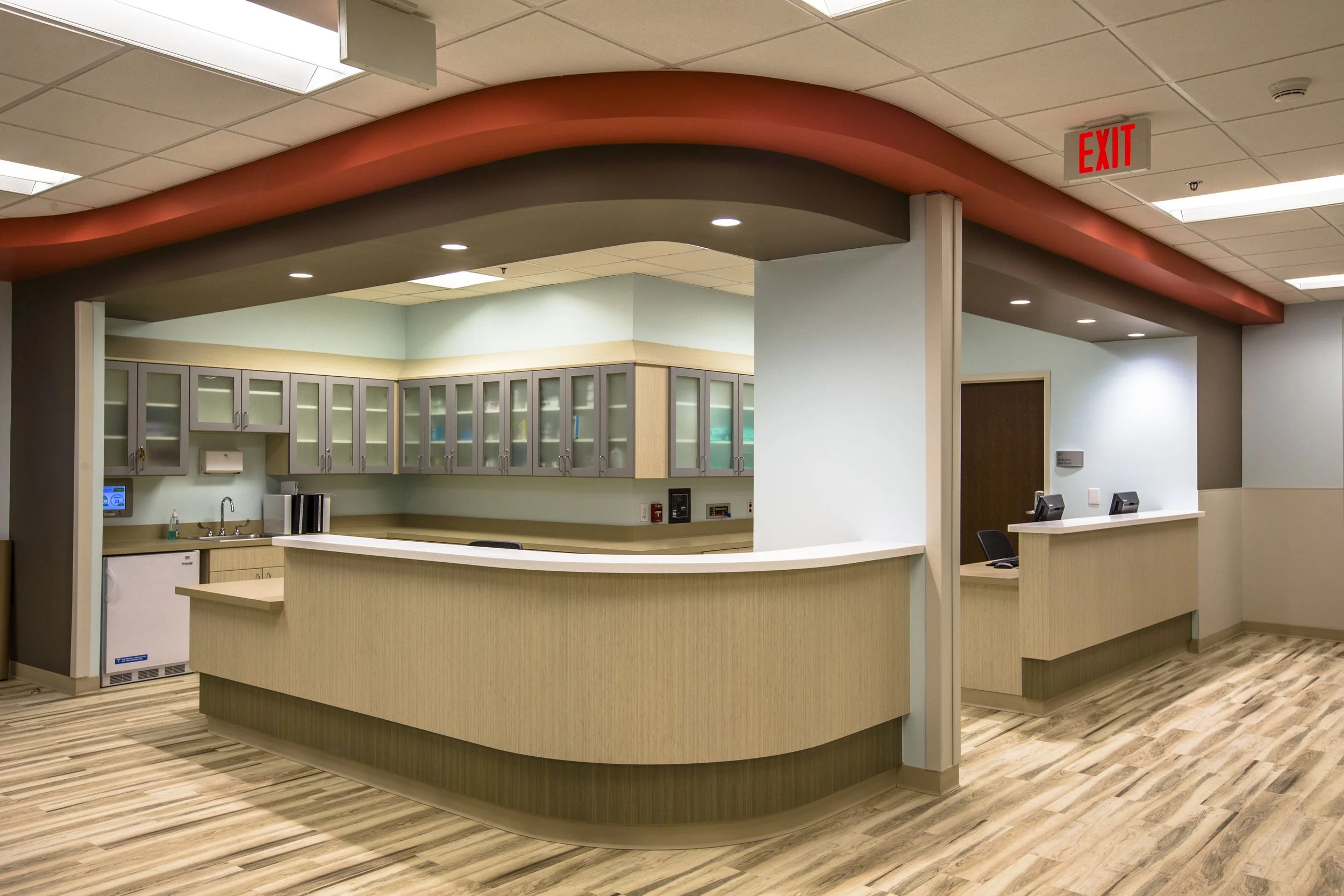 A hospital reception area with a curved wooden counter, light wood cabinets behind it, and a waiting area with chairs. An illuminated red exit sign hangs from the ceiling.