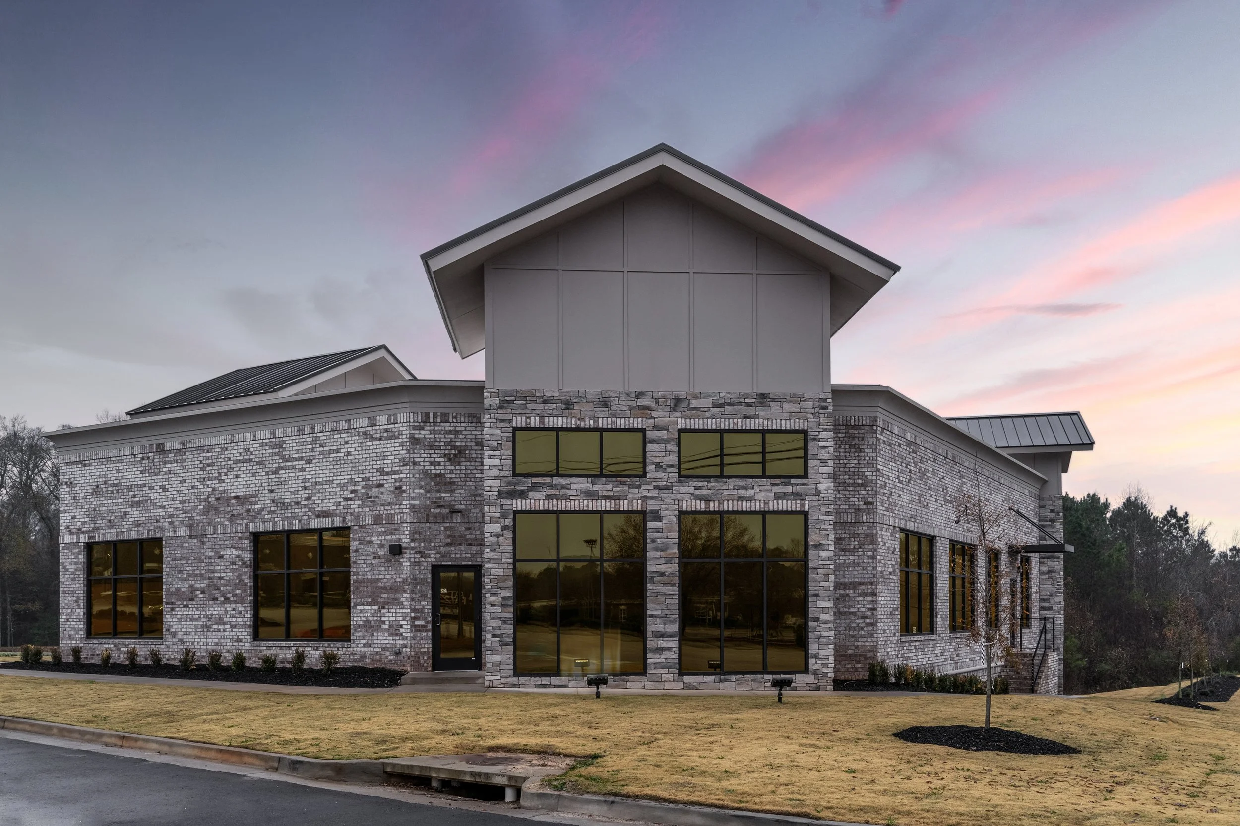 Modern brick building with large windows and a sloped roof, sunset sky with pink and purple hues, small trees and a grassy lawn in front.
