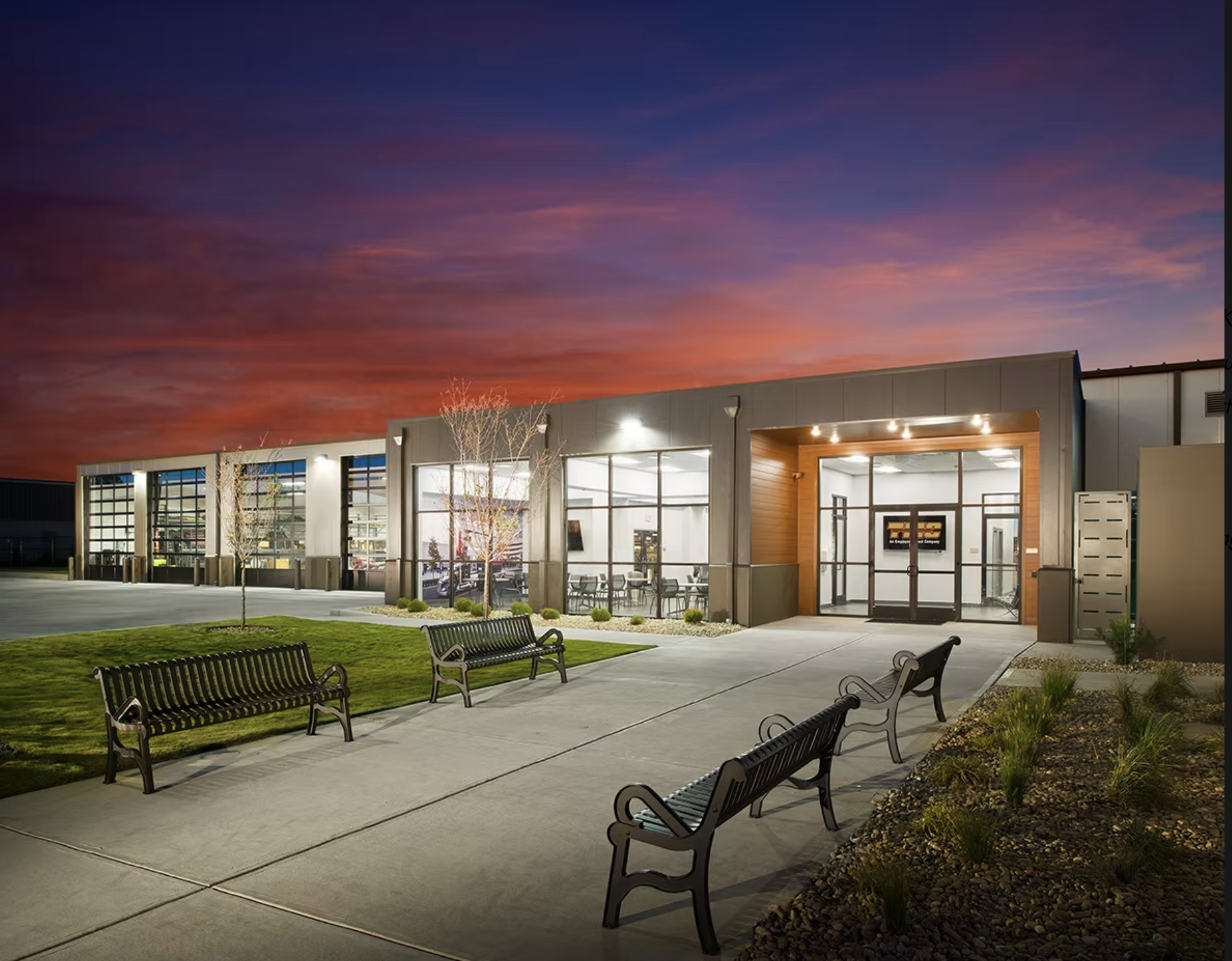 Modern building with large glass windows and a wooden entryway, surrounded by benches, trees, and landscaped bushes, under a colorful sunset sky.