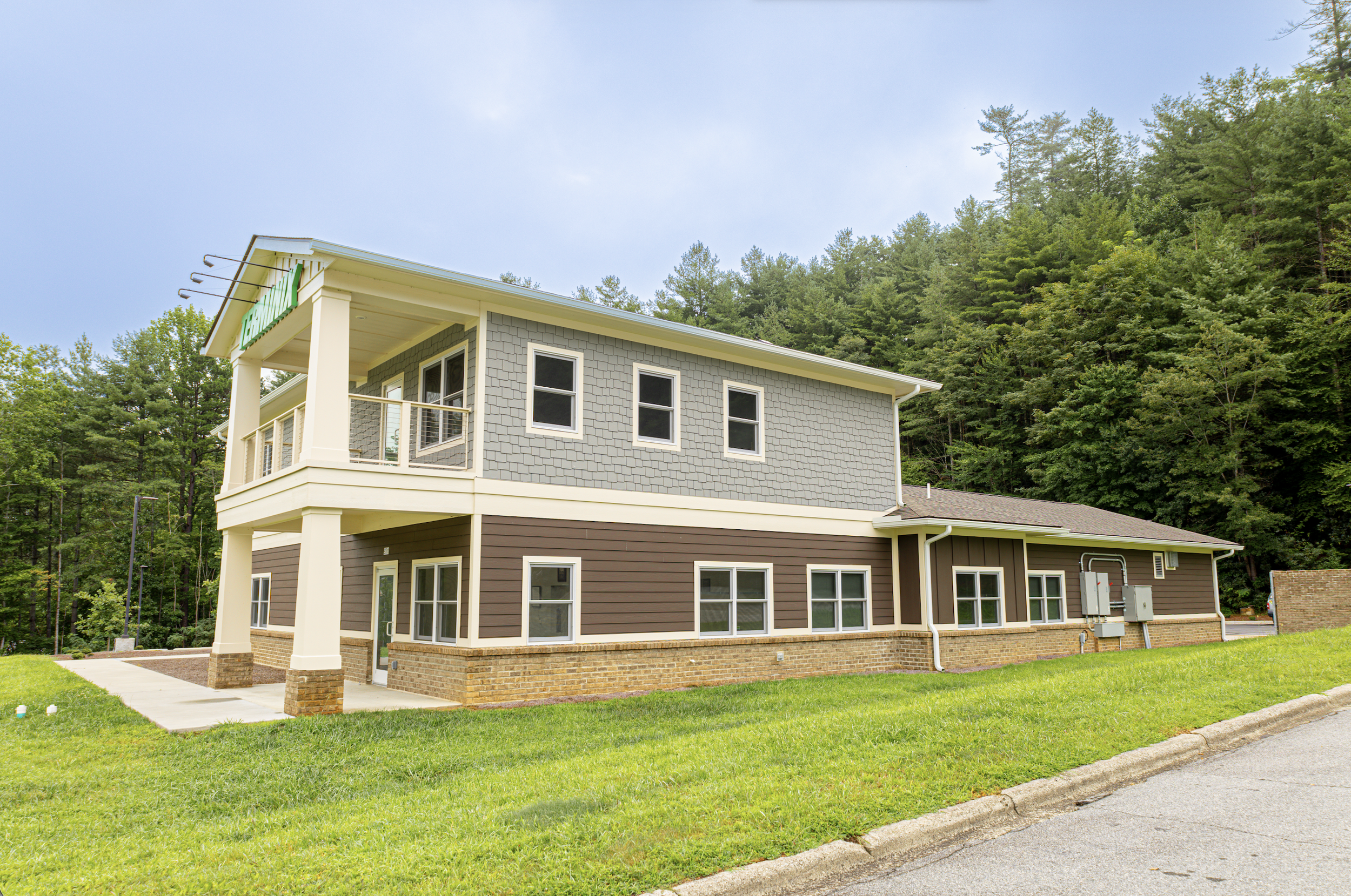 A modern multi-story house with gray and brown siding, white trim, and a brick foundation, surrounded by green grass and trees under a partly cloudy sky.