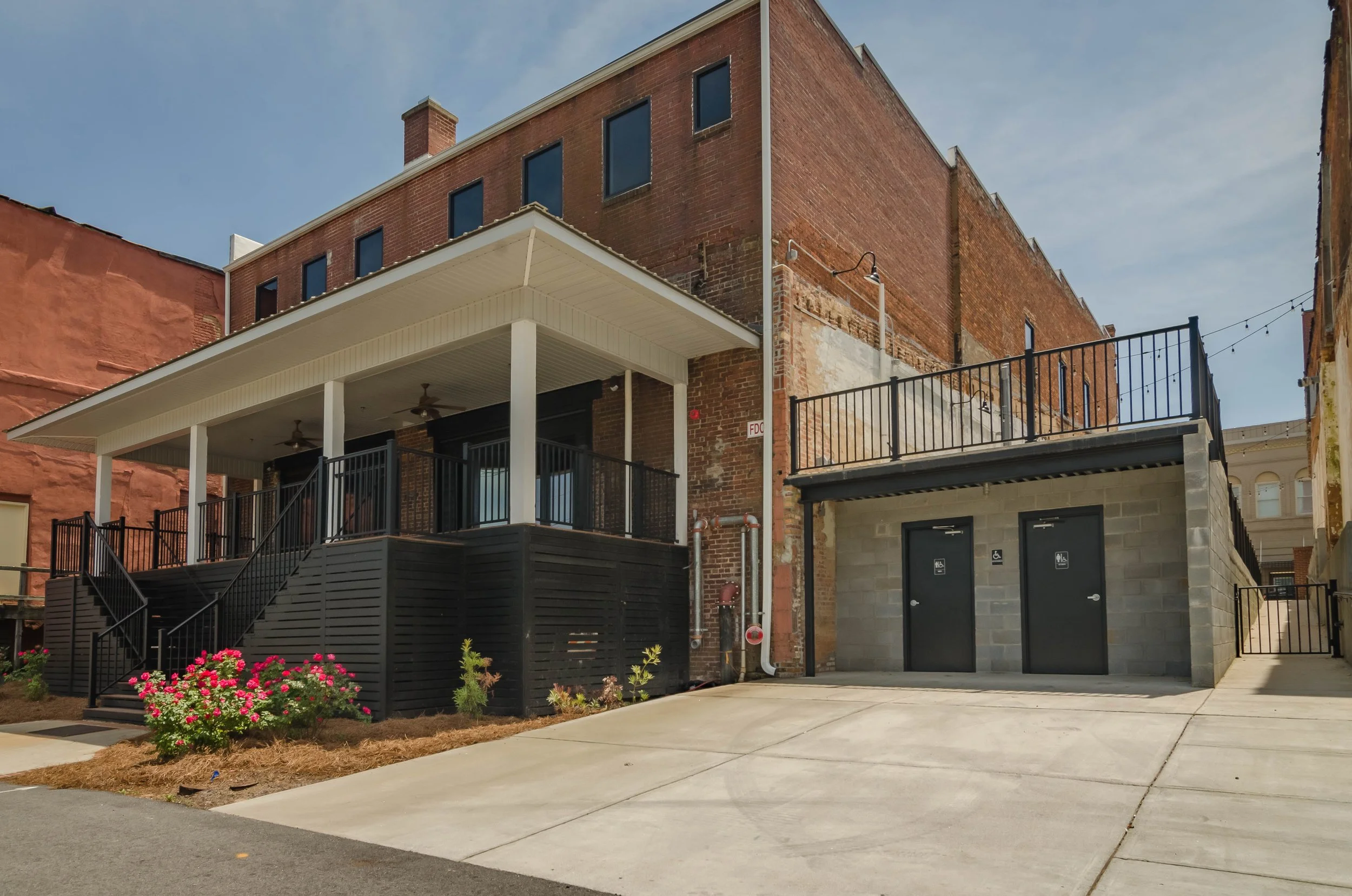 A multi-story brick building with a white porch and black railing, featuring a small garden with pink flowers in the foreground, and a concrete sidewalk and driveway leading to black doors with accessibility signs.