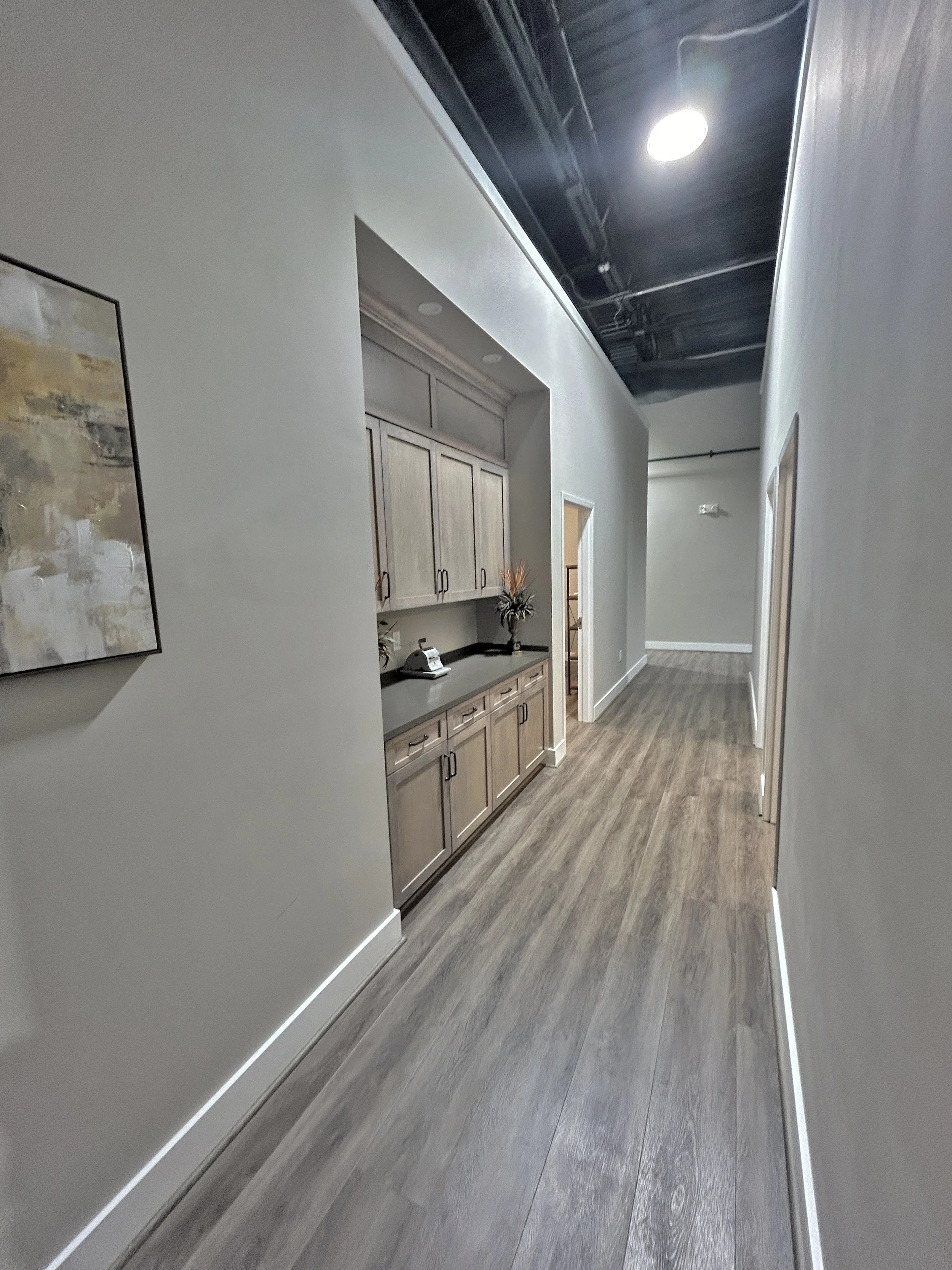Hallway with wood flooring, adjoining a room with kitchen cabinetry, and modern ceiling with black finish and recessed lighting.