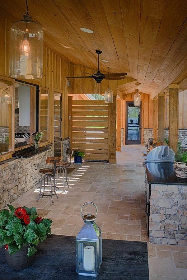 Outdoor kitchen and dining area with stone and wood accents, ceiling fan, hanging pendant lights, bar stools, potted plants, and a grill.