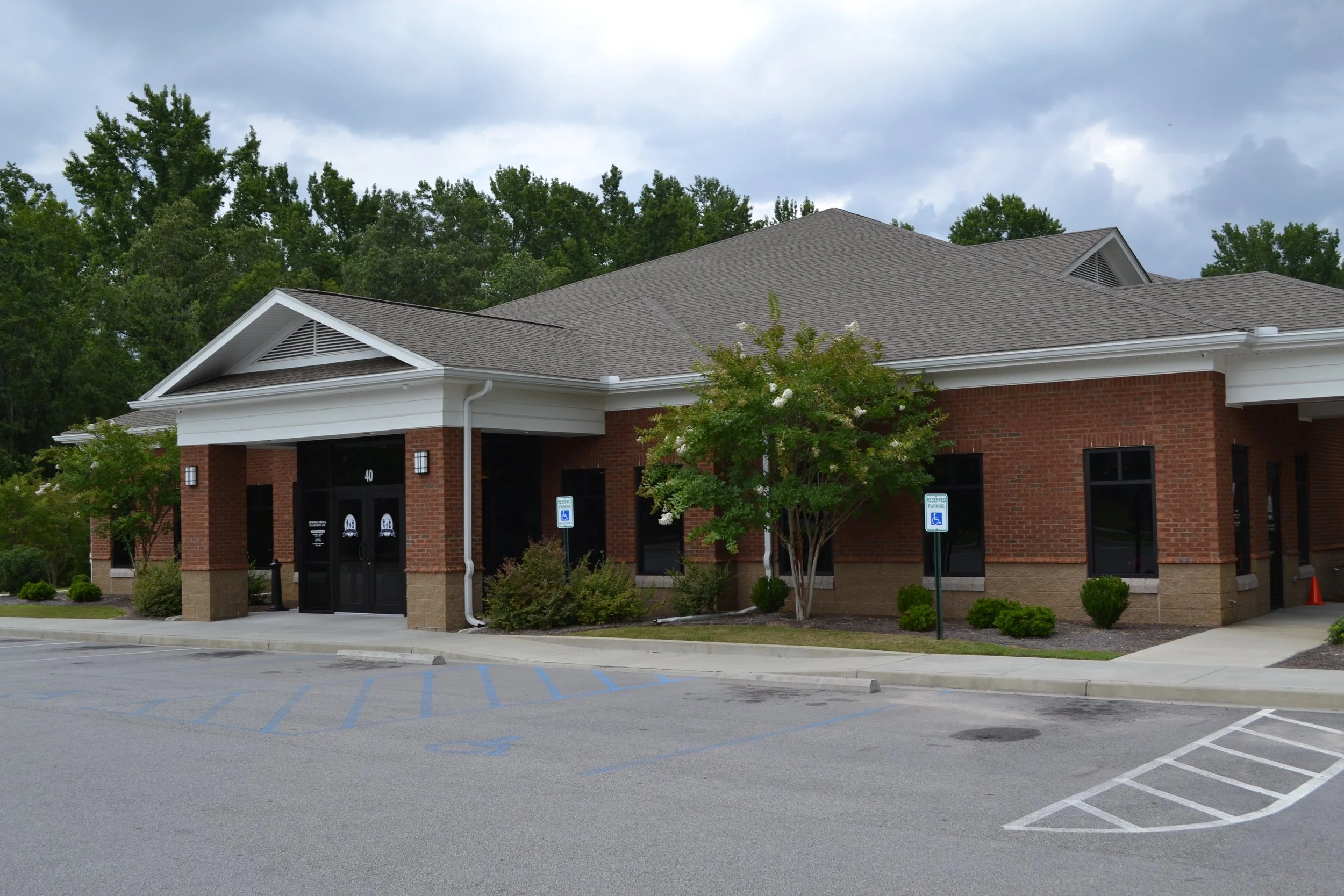 A brick commercial building with a gray shingle roof, surrounded by trees and a parking lot with designated handicapped spots and a few empty parking spaces.