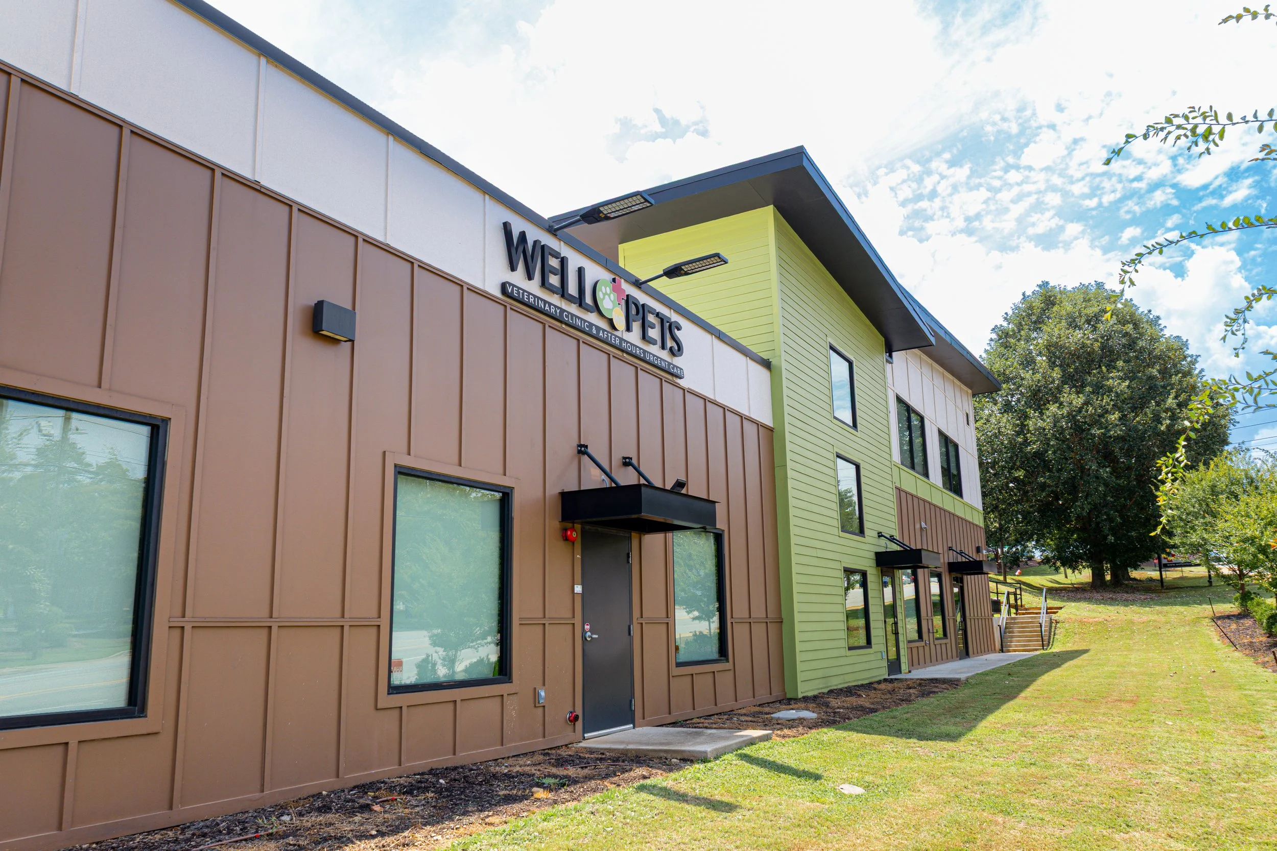 Exterior view of a modern, multi-story pet veterinary clinic with a colorful design, large windows, and a sign that reads 'Welc Pets veterinary clinic & after hours urgent care,' under a partly cloudy sky with greenery.