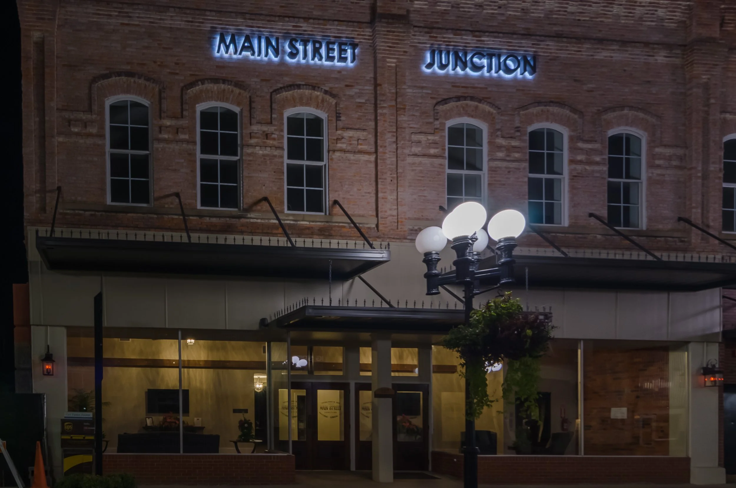 Night view of a brick building with illuminated blue neon signs reading 'Main Street' and 'Junction' on the upper floor. There are tall windows with white frames, a streetlamp with five white spherical lights, and some trees and a ground-level entrance visible in the foreground.