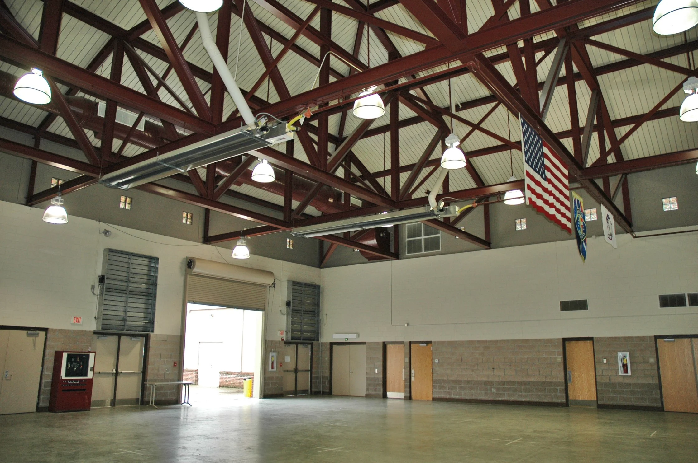 An empty indoor gymnasium or sports hall with a high ceiling featuring exposed red metal beams and several hanging white lights. The walls are white with small windows near the ceiling, and there are doors, garage-style doors, and an American flag hanging from the ceiling.