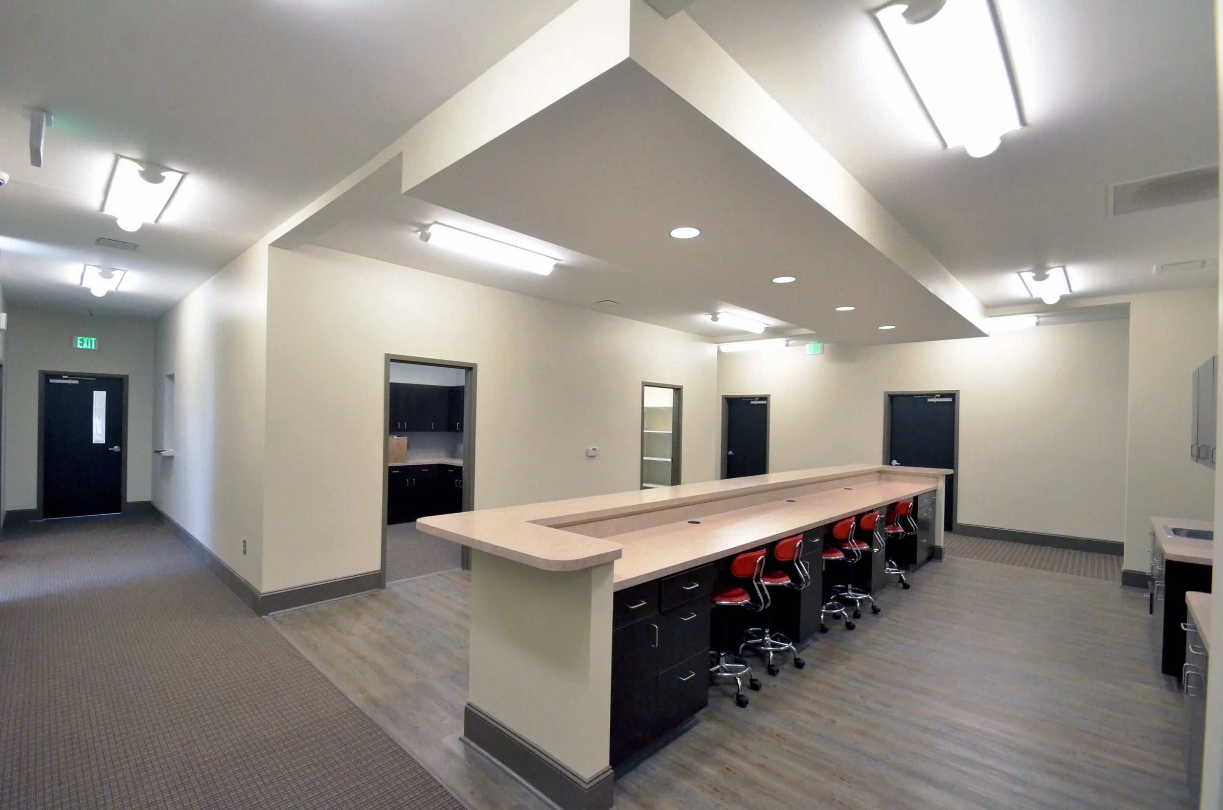 Interior of a modern room with a long counter and red chairs, multiple ceiling lights, a door, and windows showing a kitchen area.