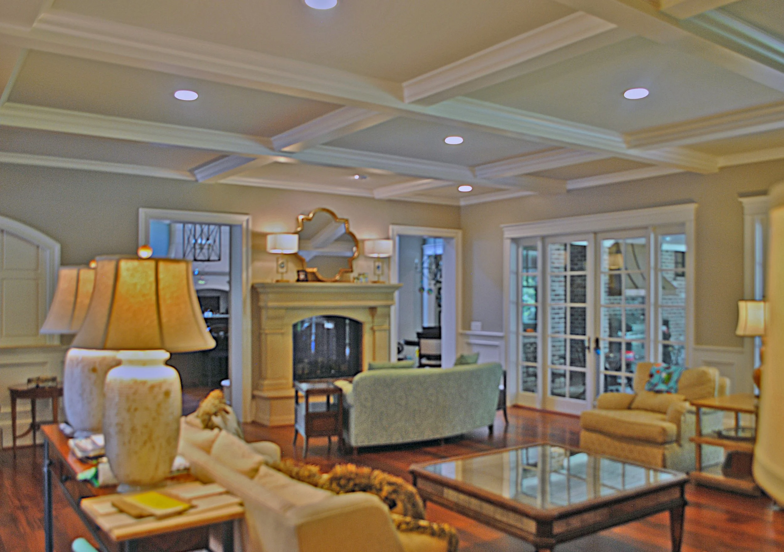 A cozy living room with a beige and green color scheme, featuring a fireplace, multiple sofas, armchairs, a glass coffee table, lamps, and large windows with white trim.