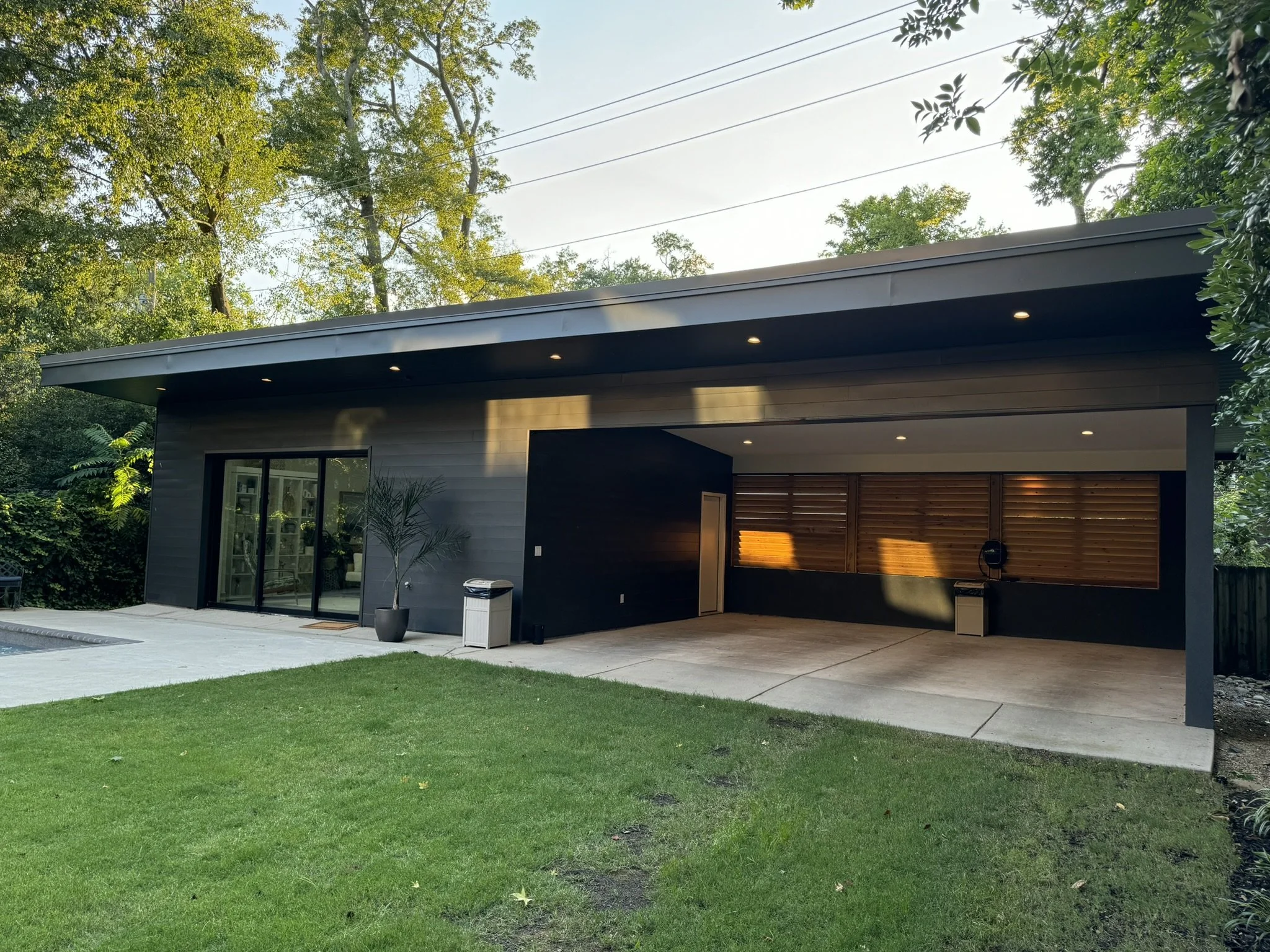 Modern house with a flat roof, large glass doors, and an outdoor patio area surrounded by trees and greenery, during sunset.