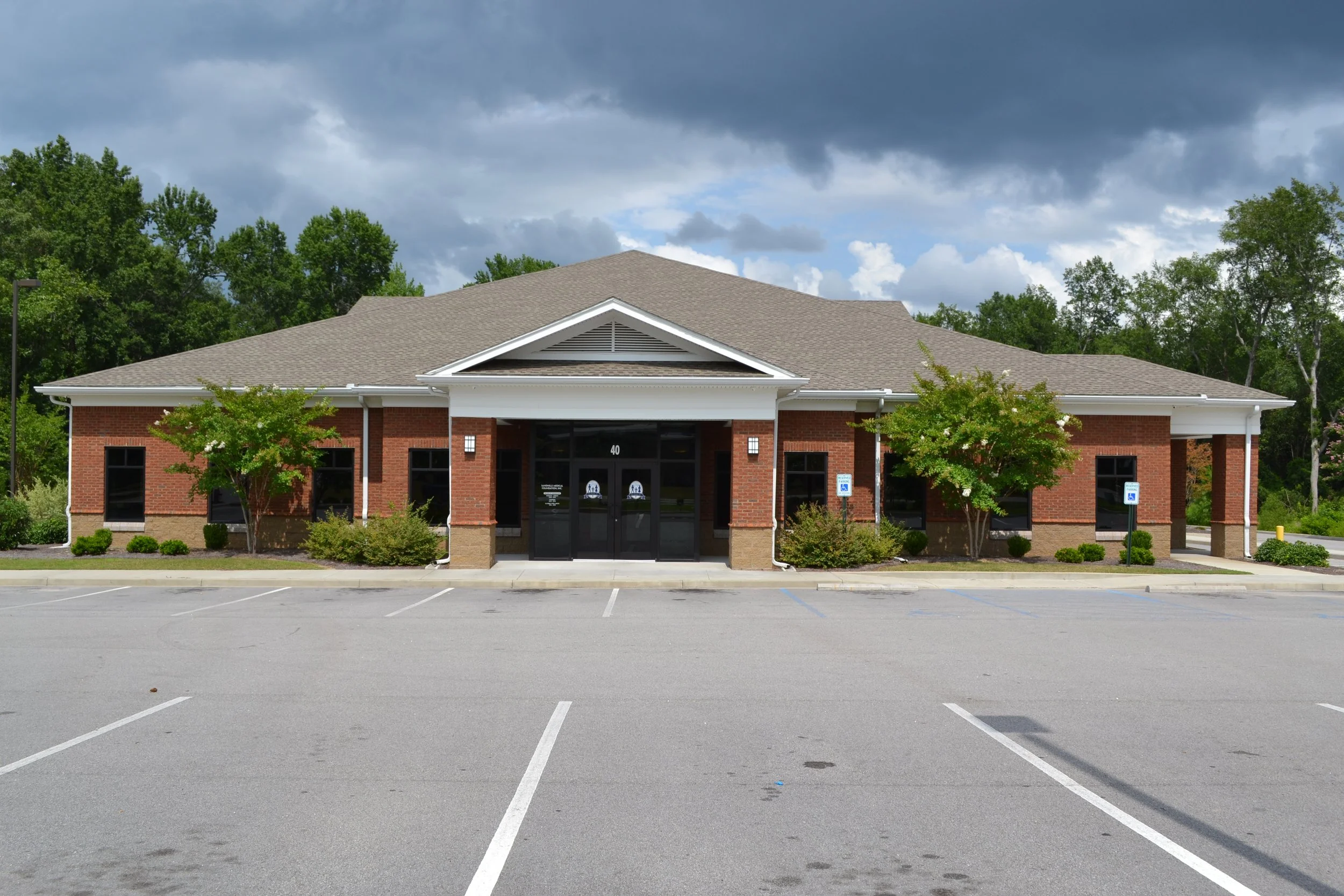 An empty parking lot in front of a brick building with trees and a cloudy sky in the background.