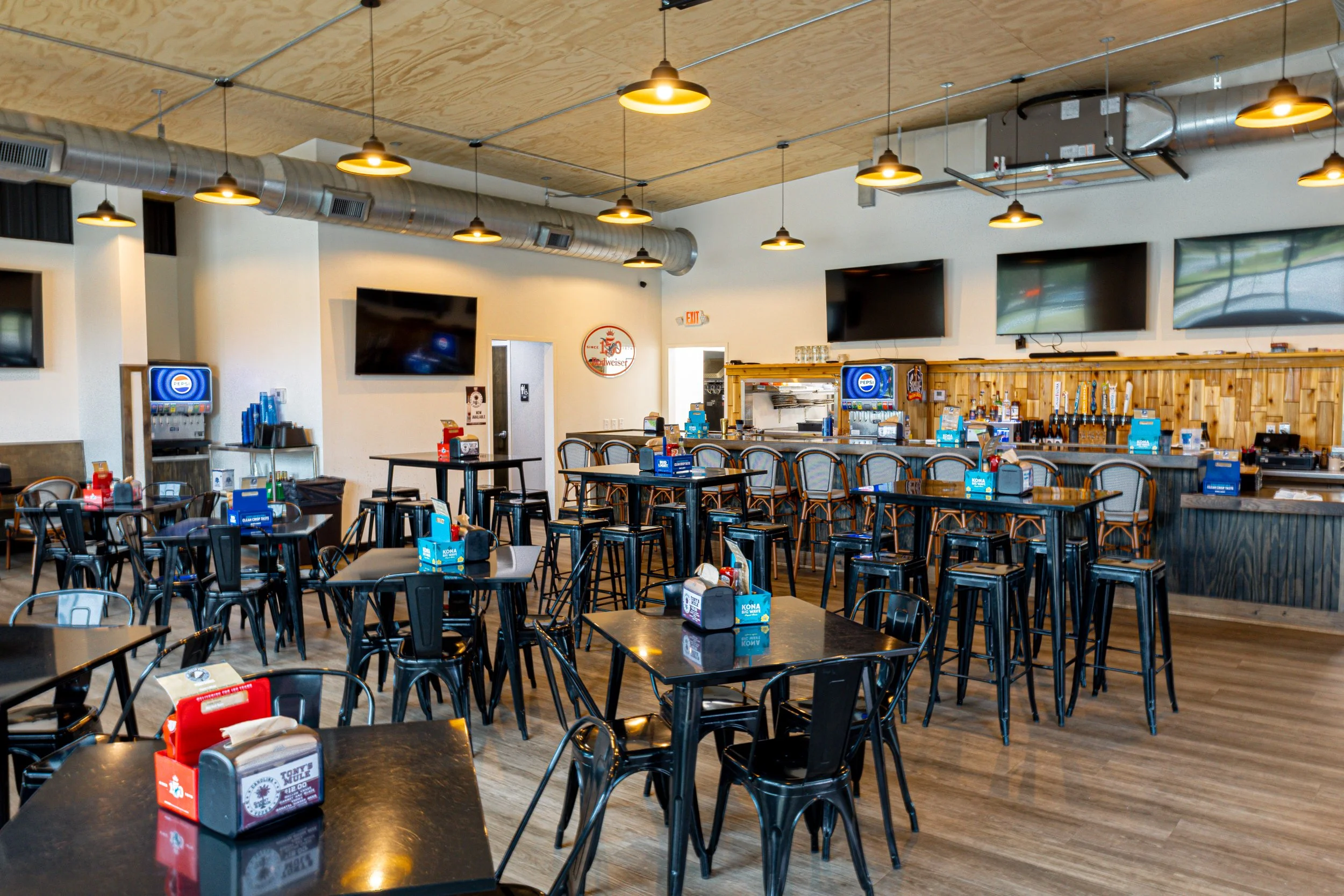 Interior of a casual restaurant with black tables, metal chairs, multiple mounted flat-screen TVs, pendant lighting, and a wooden bar area.
