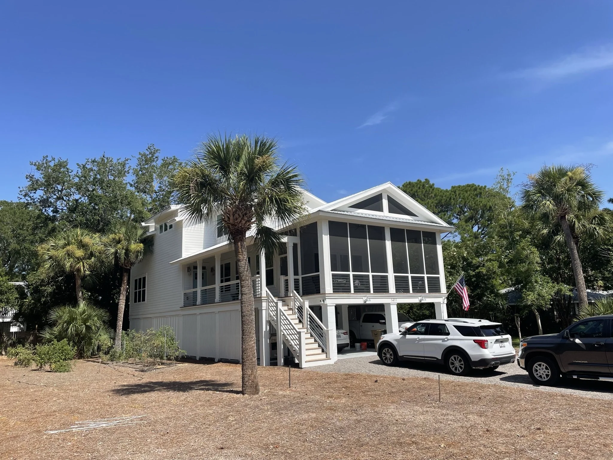 White two-story house with screened porch, steep gabled roof, and palm trees in the front yard, with several parked cars in front and a clear blue sky overhead.