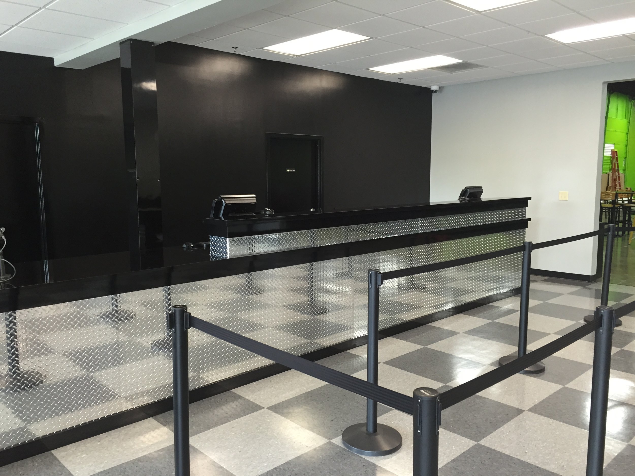 An empty check-in or reception counter in an indoor setting with a black wall and metallic surface, surrounded by black queue barriers. There is a small room or door behind the counter and a green wall with furniture visible through an open doorway o