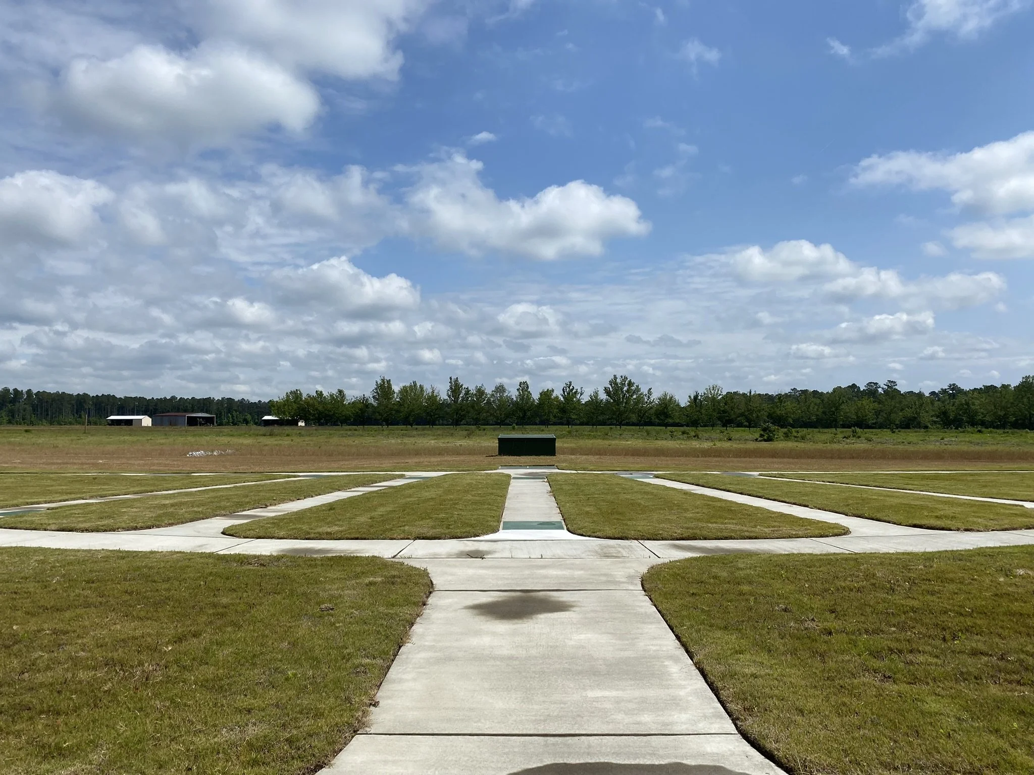 An outdoor scene with a concrete path leading to a grassy field, trees in the background, and a partially cloudy blue sky.