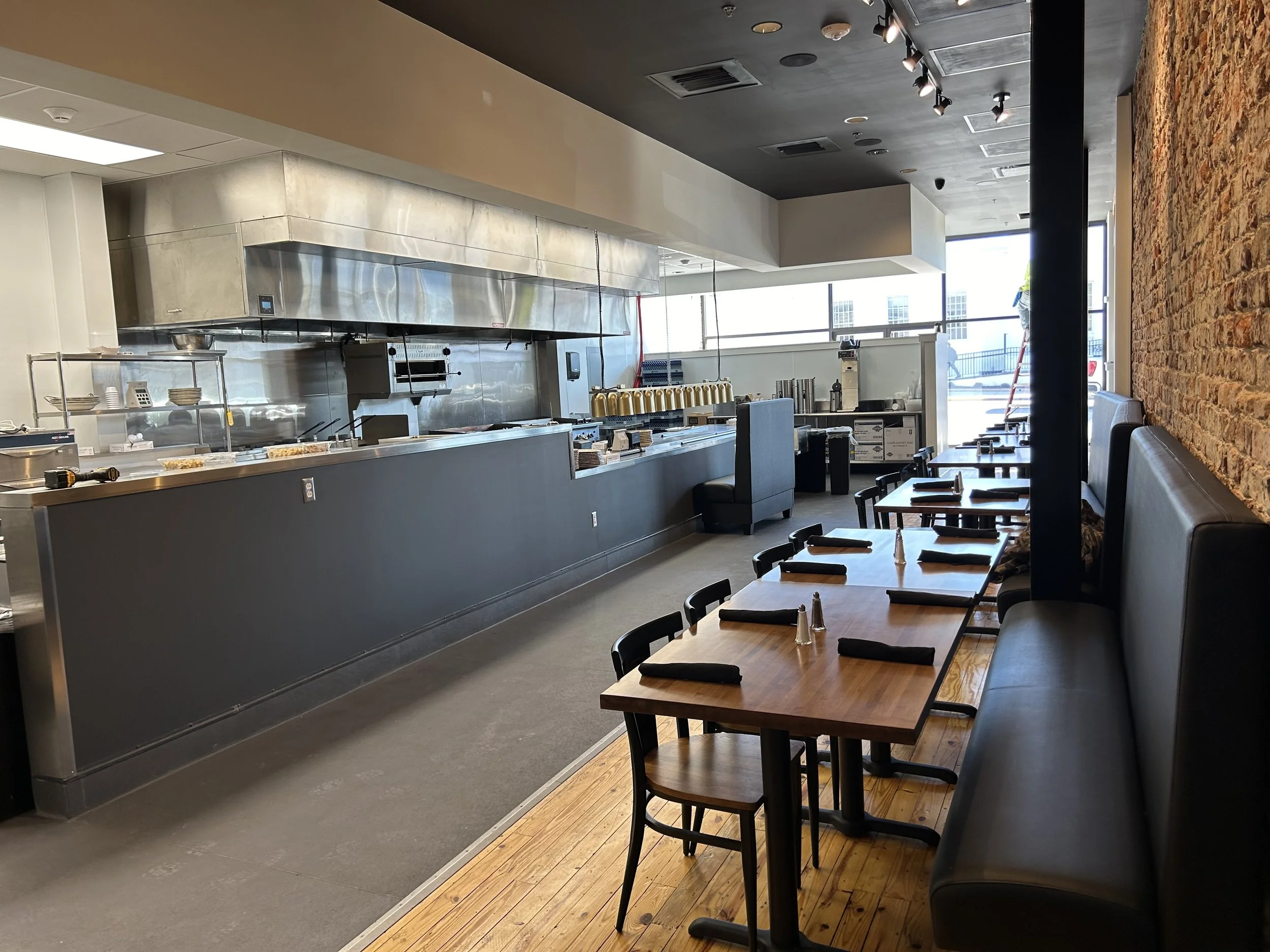 Empty restaurant interior with tables, chairs, exposed brick wall, and large windows letting in natural light.
