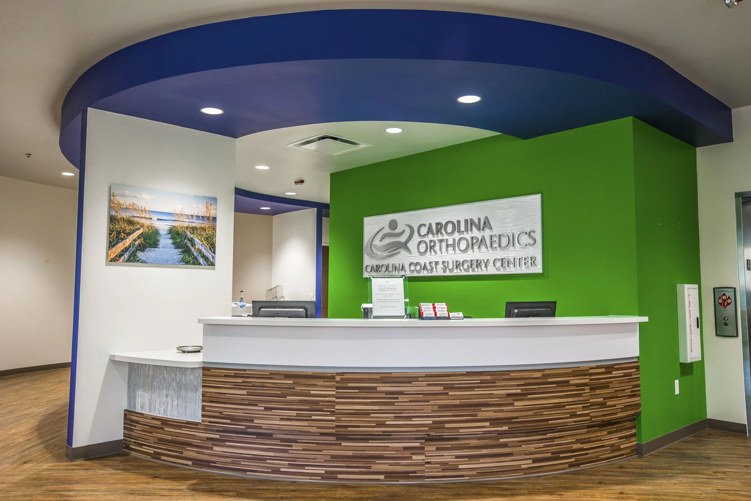 Reception desk at Carolina Orthopaedics in a modern, brightly lit medical office with green accent wall, a wooden panel front on the desk, and a landscape photograph of a beach with sand dunes and vegetation on the white wall behind.