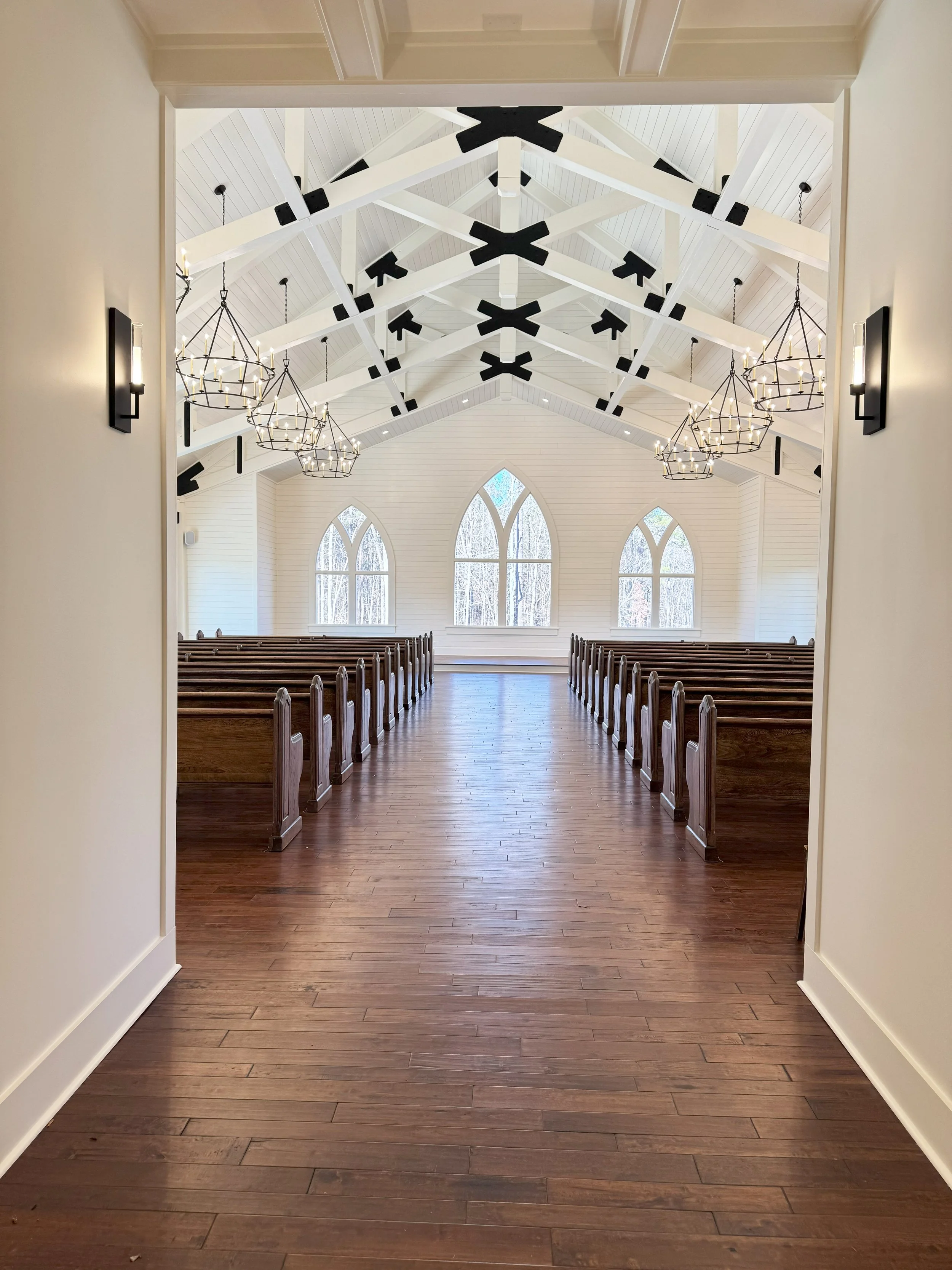 View of the interior of a church with wooden pews, large arched windows, white walls, chandelier-style lights, and exposed white ceiling beams with black accents.