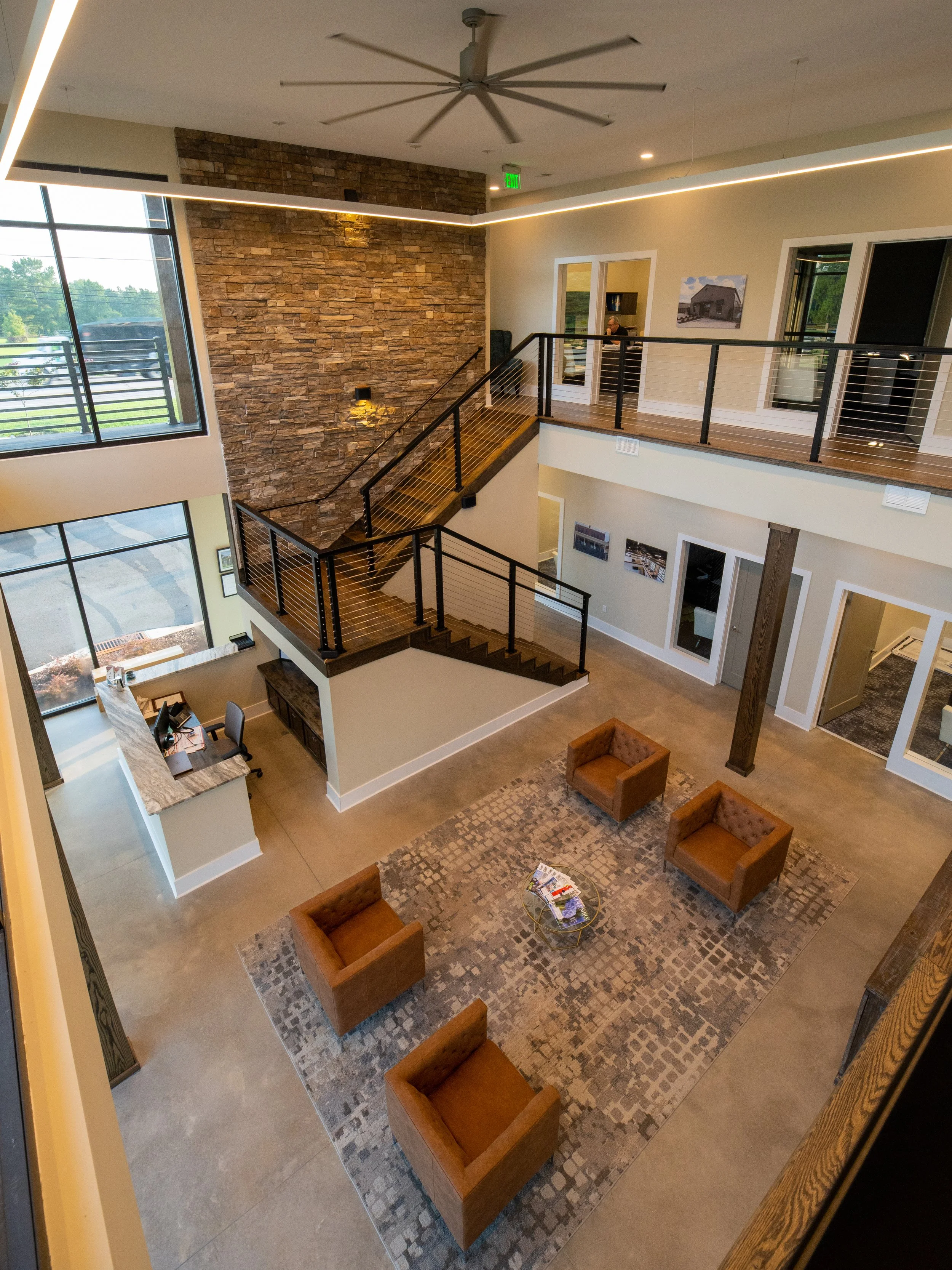 Interior of a modern building with an open second-floor view overlooking a seating area with four brown armchairs around a small round glass coffee table, a patterned rug, and a stone accent wall with a large window to the left and several doorways o