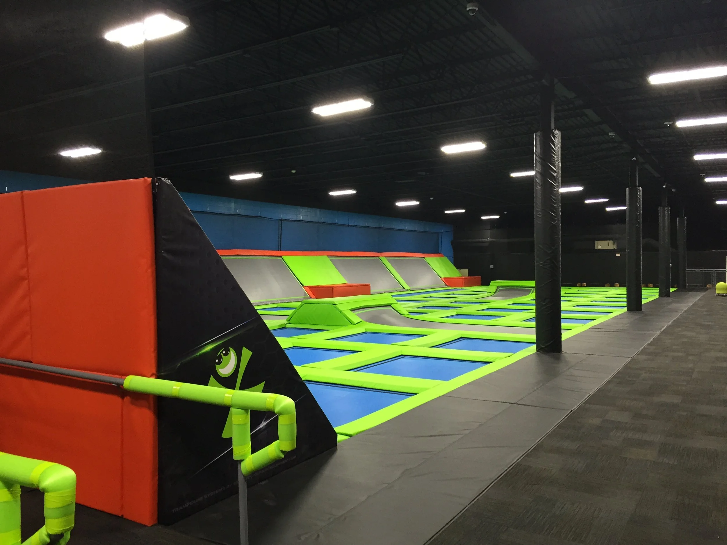 An indoor trampoline park with multiple interconnected trampolines in bright green, blue, and orange colors. The area is surrounded by black padding and safety barriers, with some black poles and overhead lighting visible.