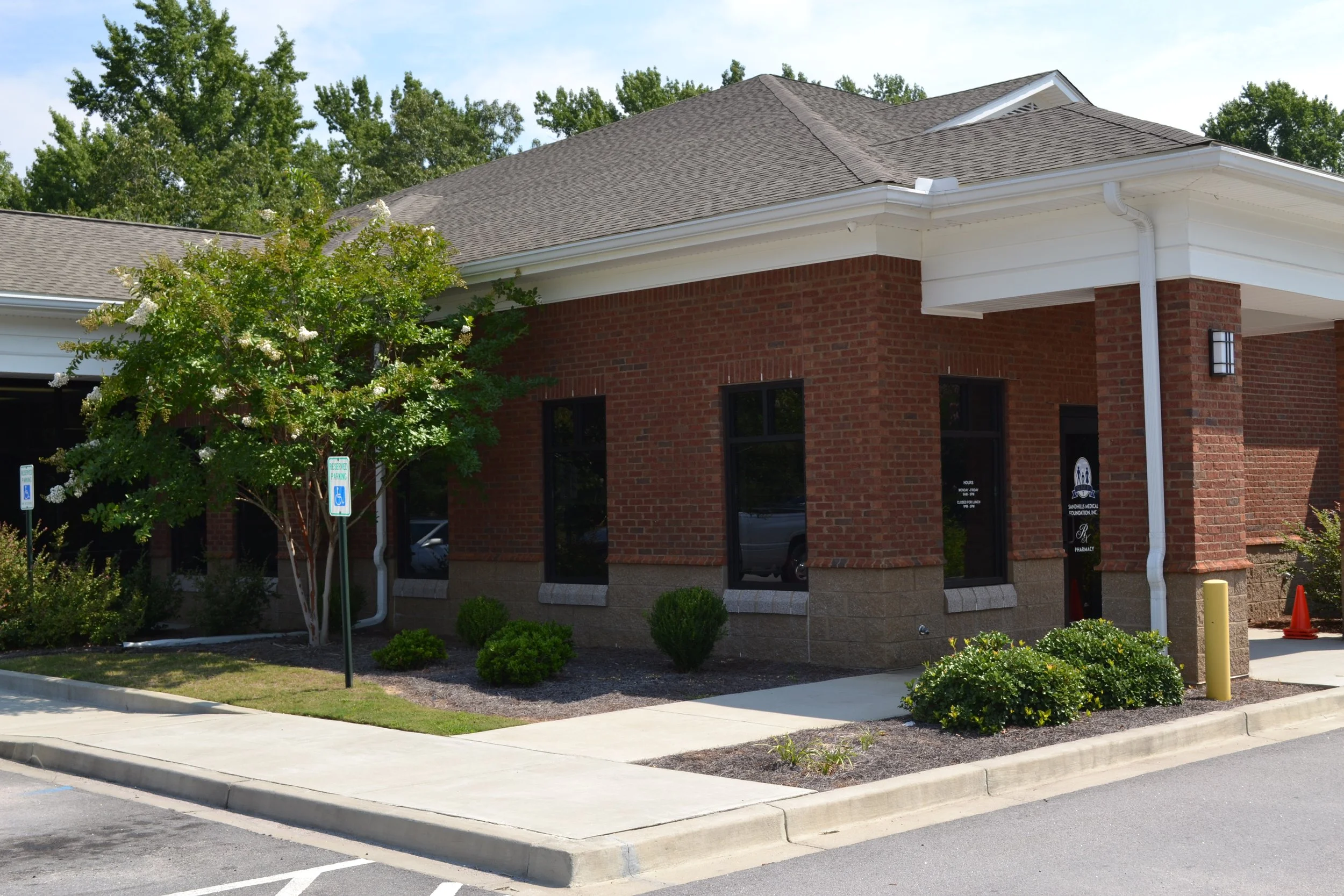 A brick building with a gray shingled roof, large windows, a small landscaped area with bushes and a tree, and parking signs indicating reserved parking spots.