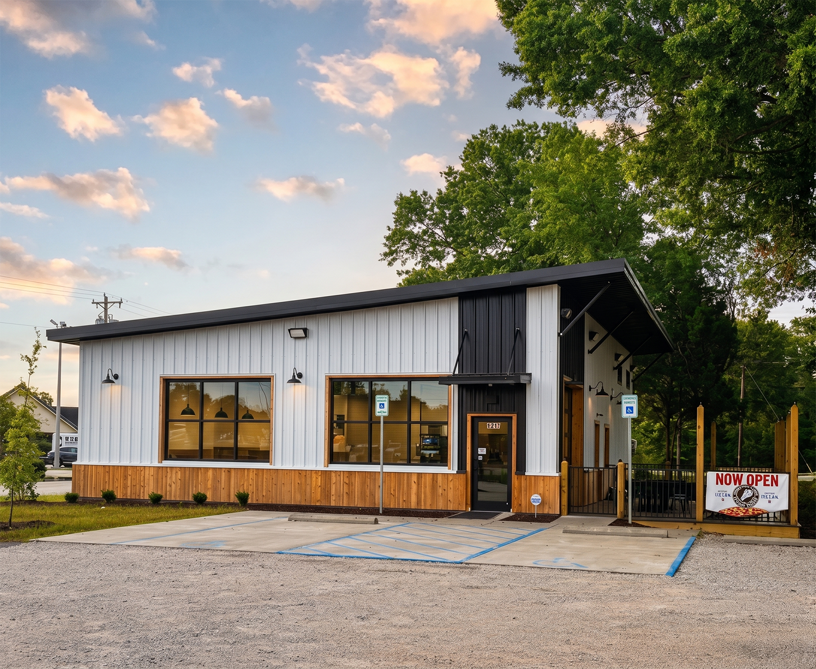 Newly opened modern restaurant building with large windows, black and white exterior, wooden accents, and a sign reading "NOW OPEN". Parking spaces are marked in front, and there are handicapped parking signs near the entrance. The building is surrounded by trees and a partly cloudy sky.
