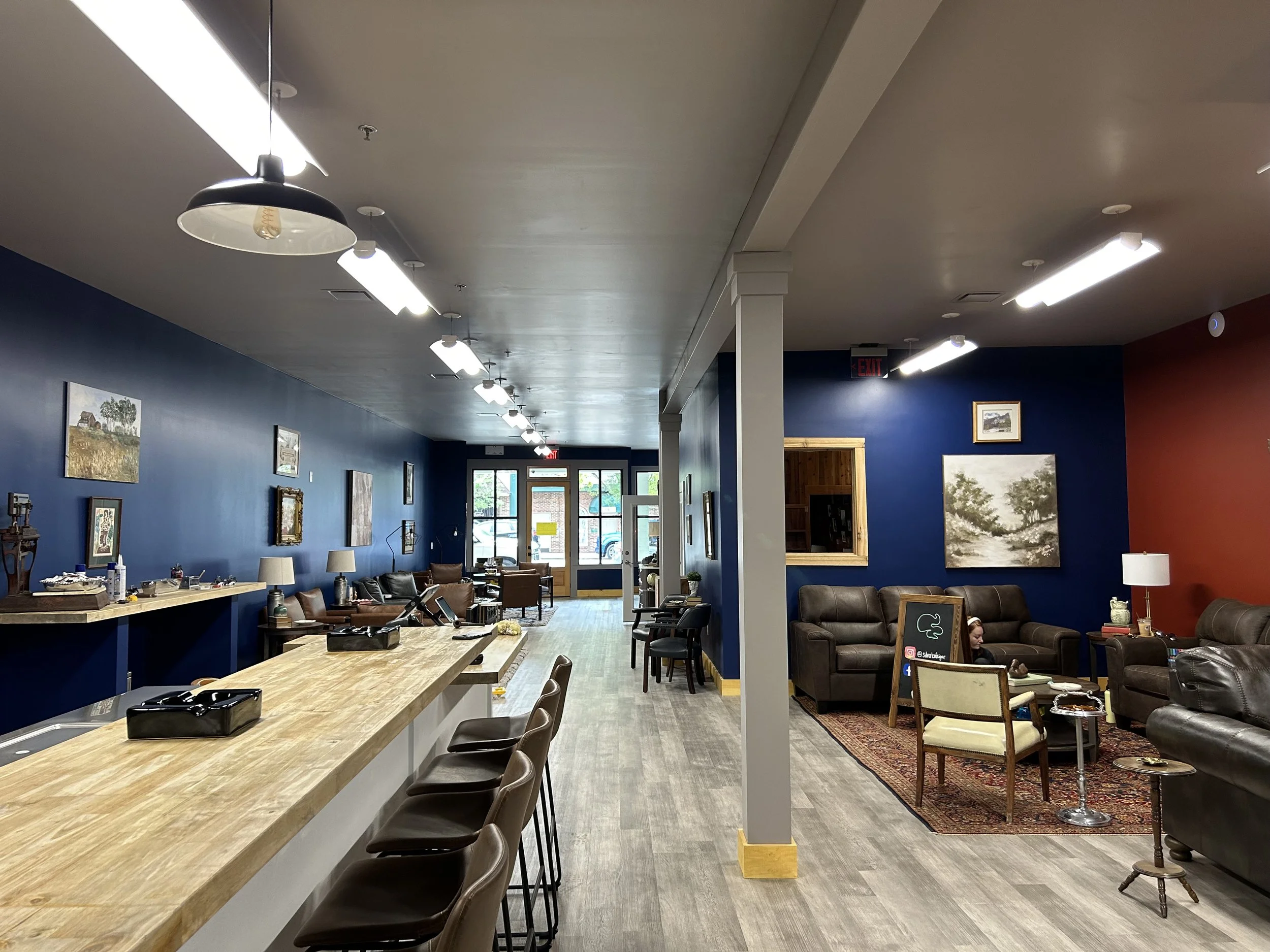 A spacious interior with a long wooden counter on the left, brown chairs, and a seating area with sofas and armchairs on the right. The walls are painted dark blue and red, decorated with framed landscape paintings. Bright overhead lights illuminate 