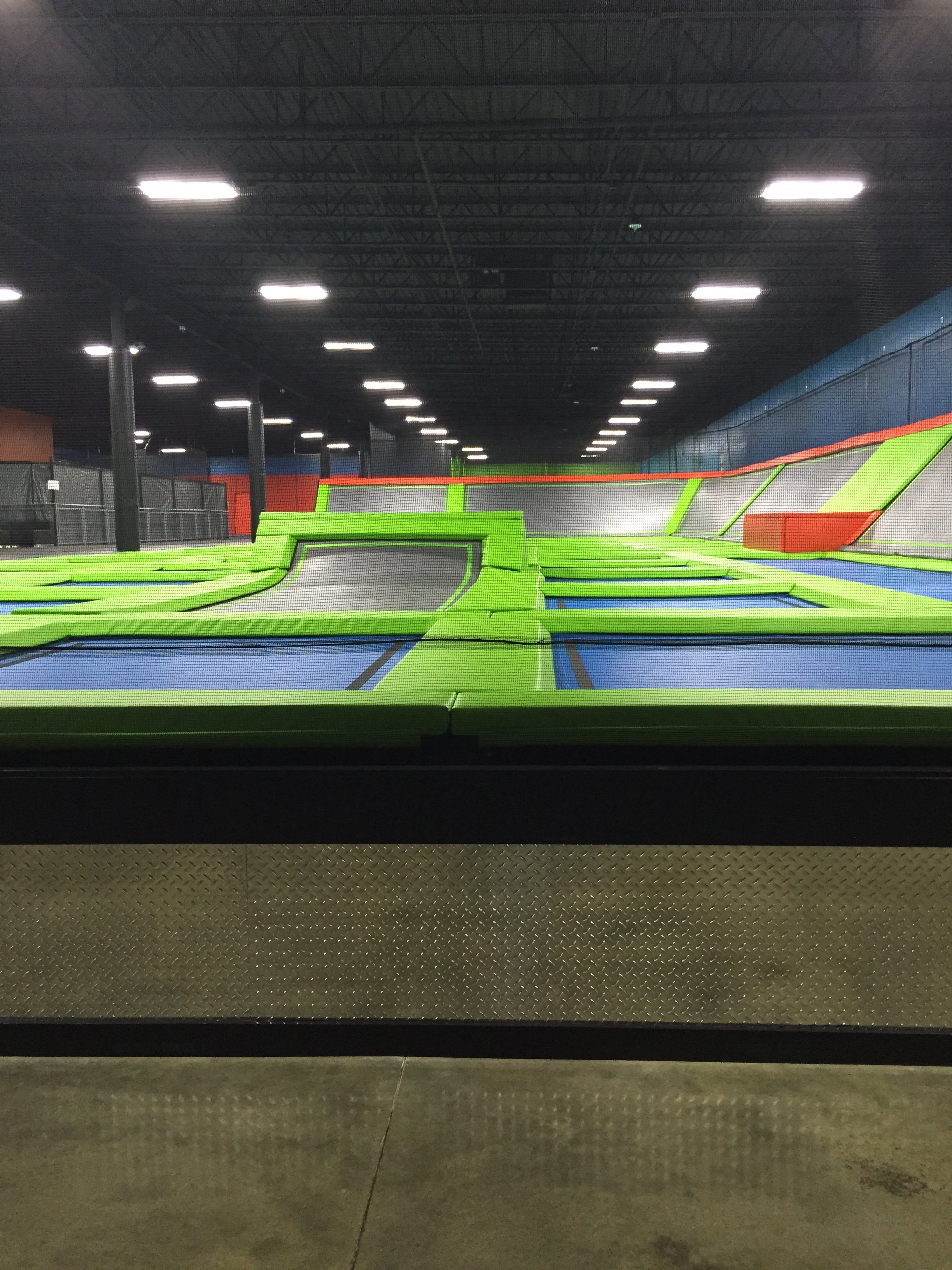 Indoor trampoline park with neon green, blue, gray, and orange trampolines and padding, illuminated by multiple ceiling lights, with black ceiling beams and a chain-link fenced area in the background.