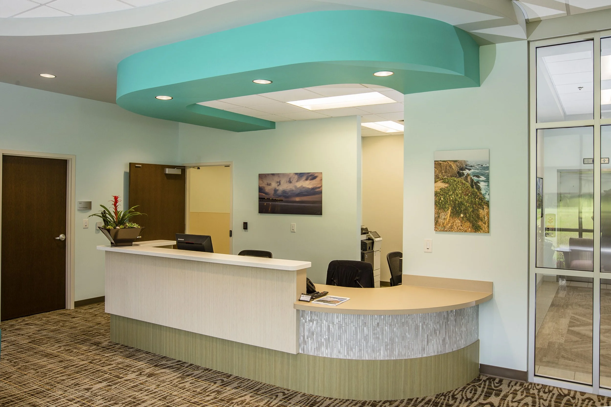 Office reception area with curved white reception desk, mint green walls, wall-mounted art, ceiling with green accent design and recessed lighting, potted plant, chairs, and a glass door.
