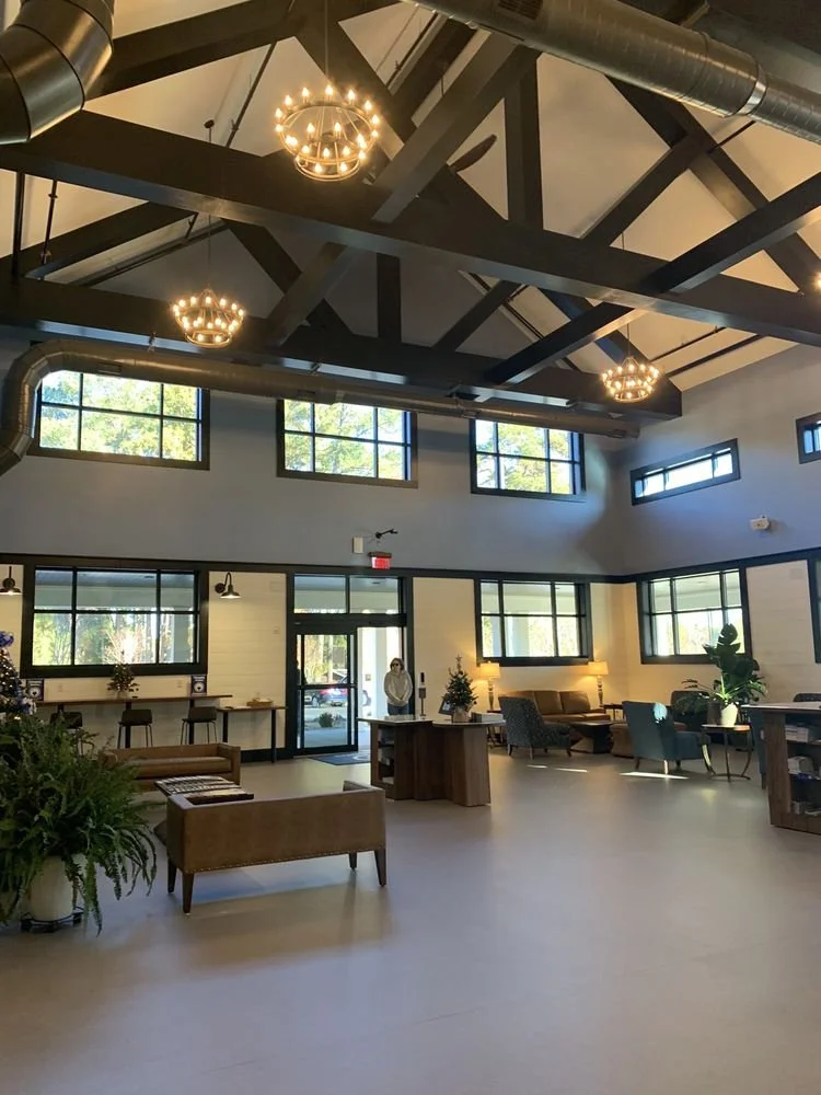 Interior of a modern, spacious lobby with high ceilings, exposed beams, and large windows. There are chandeliers, seating area with couches and chairs, potted plants, and a front desk with a person standing nearby.