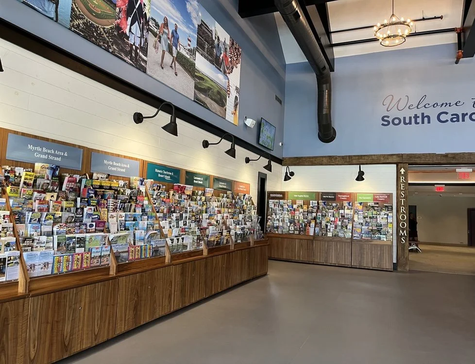 Interior of a visitor center with brochures and maps on display, including signs for Myrtle Beach and South Carolina, and a sign indicating restrooms.