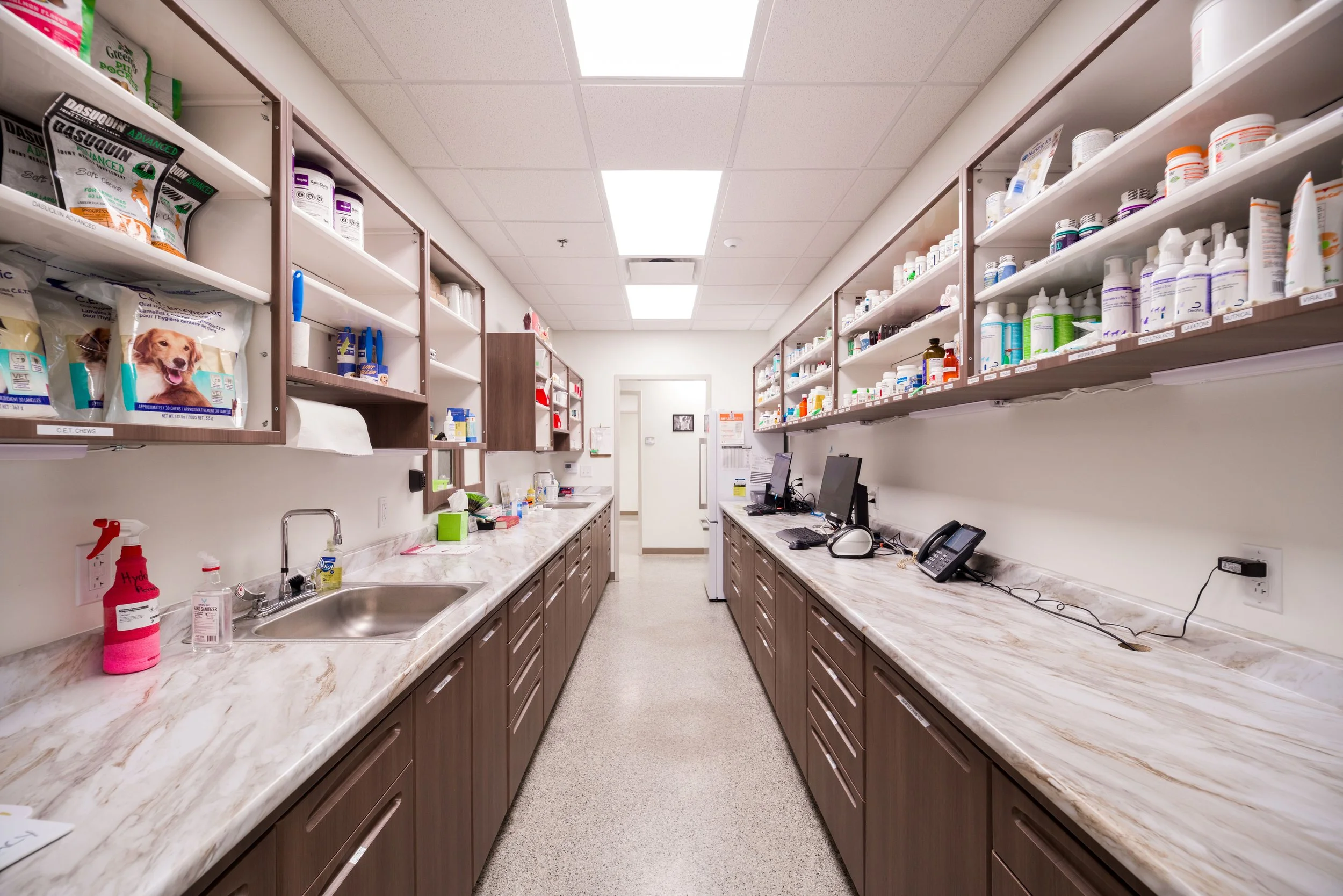 A veterinary clinic supply room with shelves stocked with medication bottles, supplies, and pet food, and a long countertop with a sink, computers, and phones.