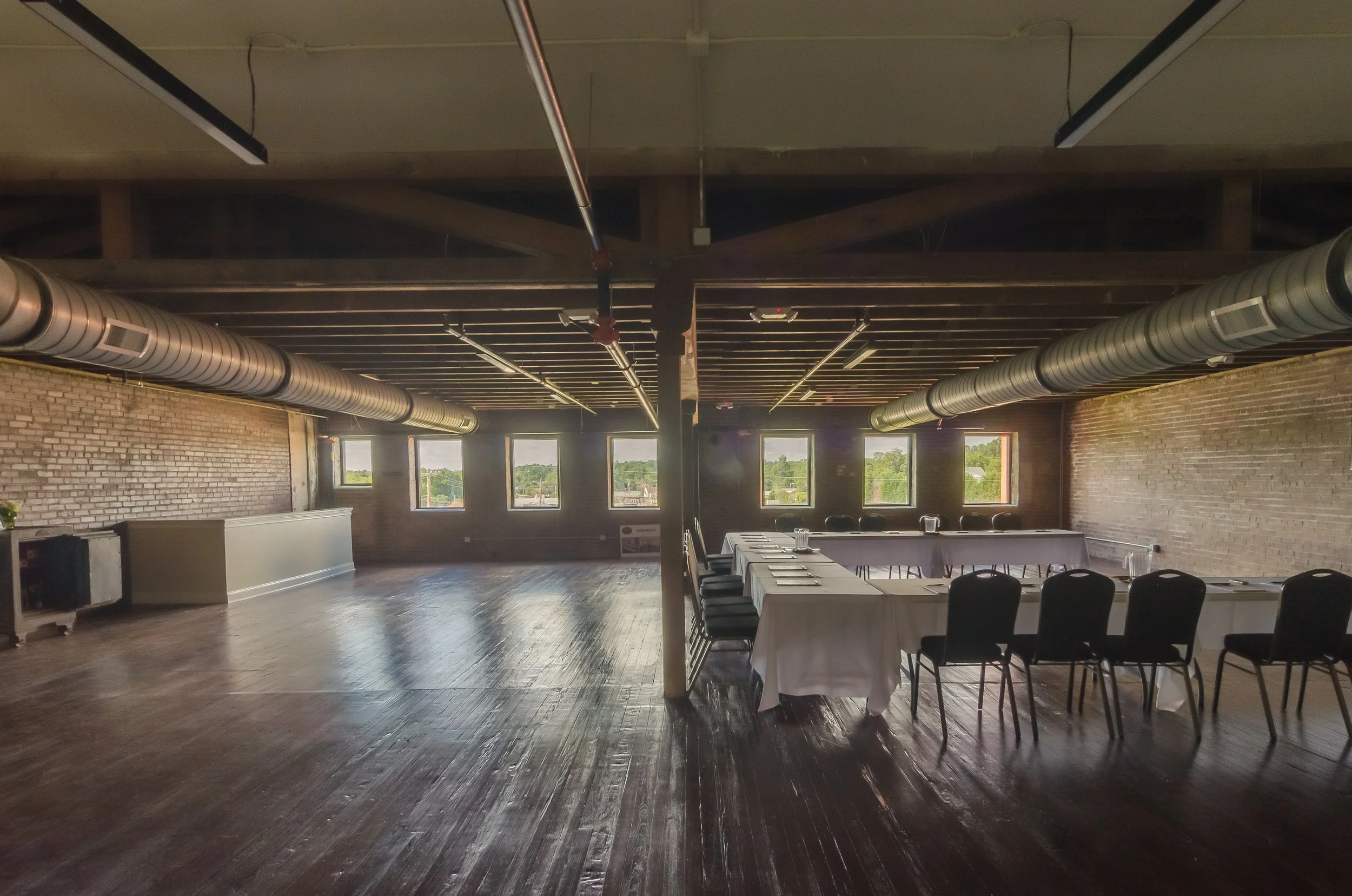 An indoor event space with a long table set for a meal, surrounded by black chairs, under a ceiling with exposed ductwork and wooden beams, with windows in the background showing trees outside.