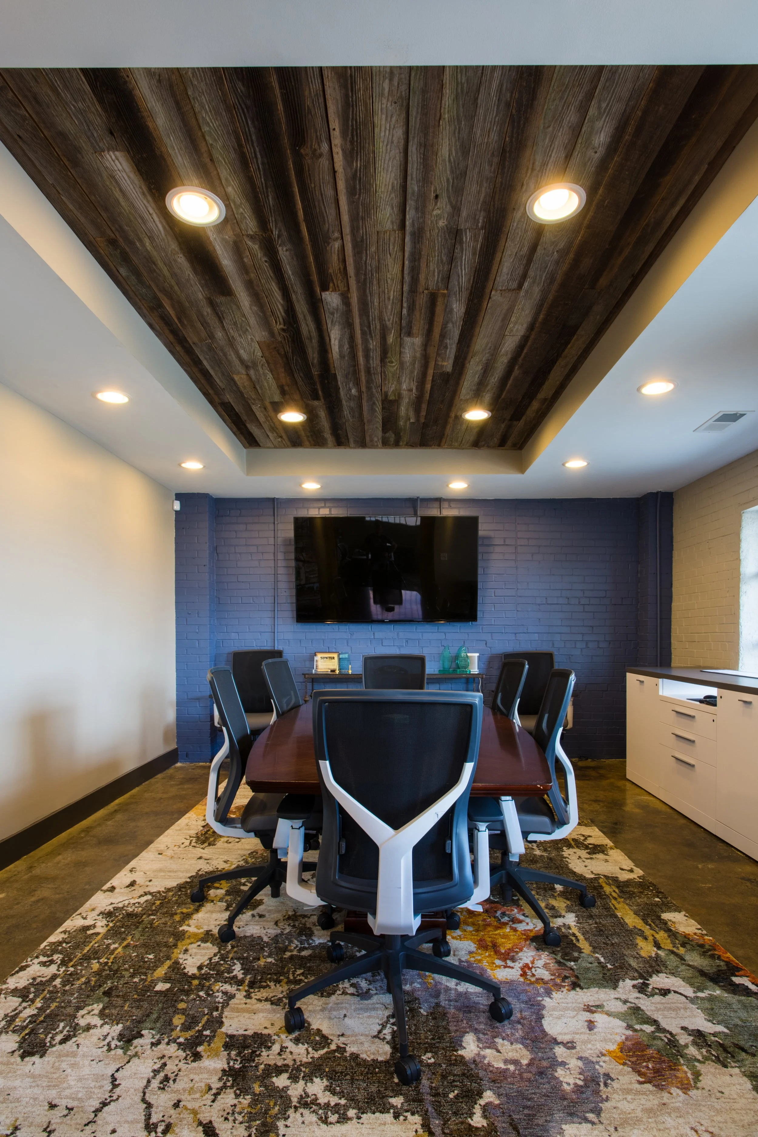 A conference room with a wooden ceiling and recessed lighting, a large TV on a blue brick wall, a table with office chairs, a carpet with a colorful abstract pattern, and a white cabinet.