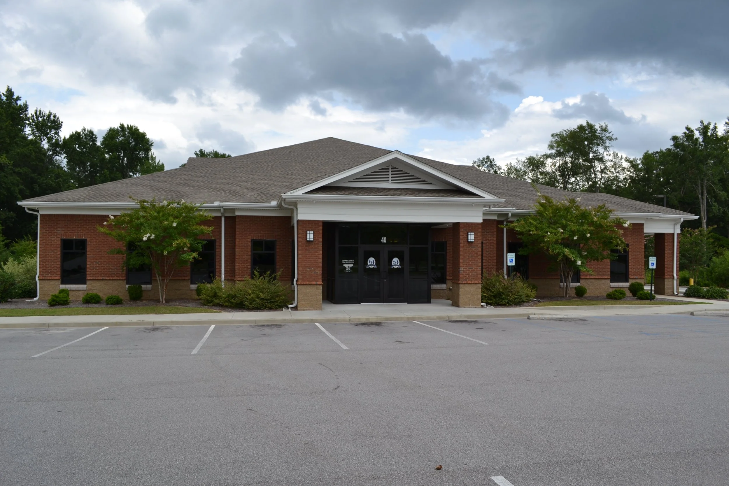 A brick building with a dark gray roof, surrounded by small trees, with an empty parking lot in front and a cloudy sky above.
