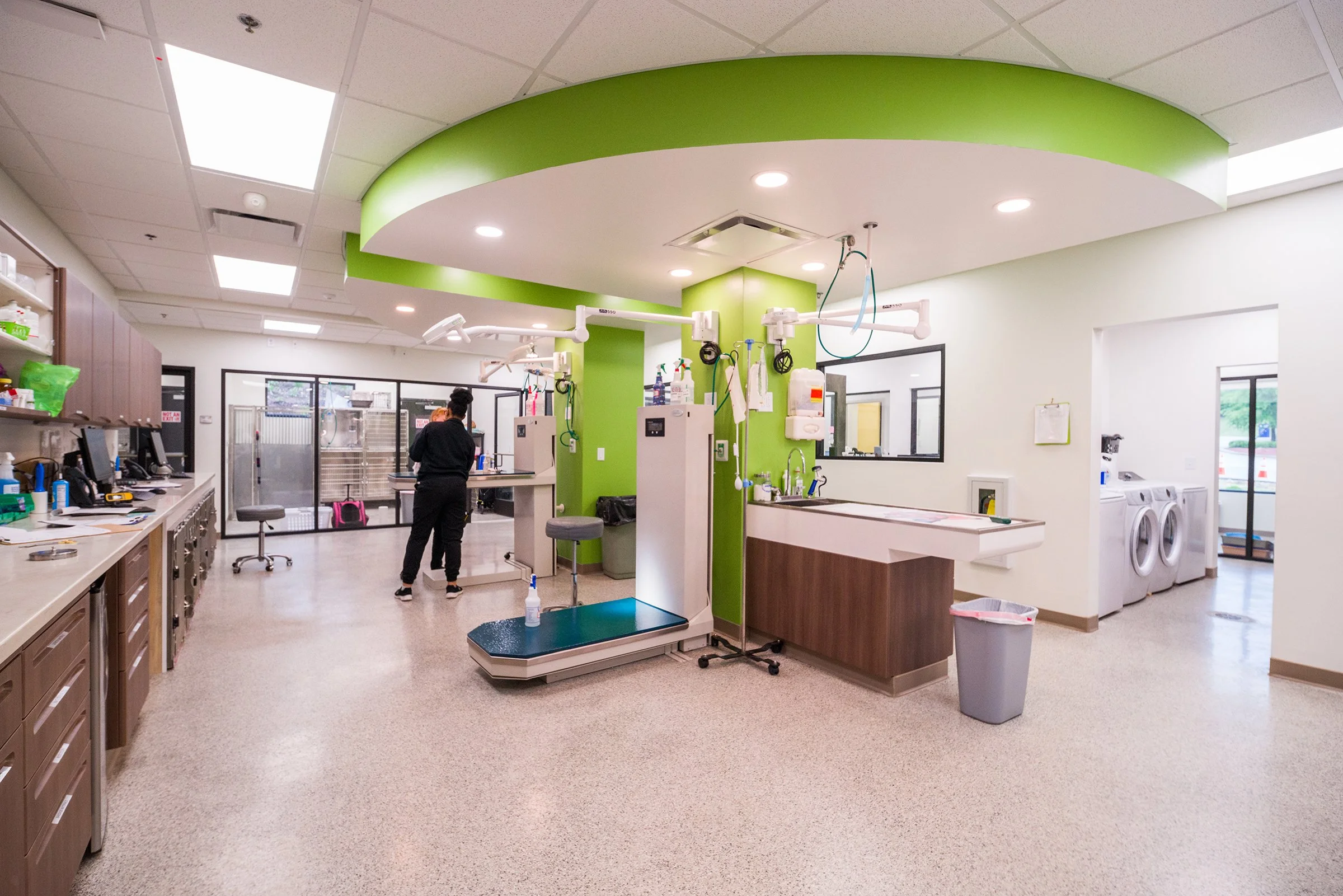 A hospital room with medical equipment, a scale, and laundry machines, with a person standing near the center, and windows showing a glimpse of the outdoors.