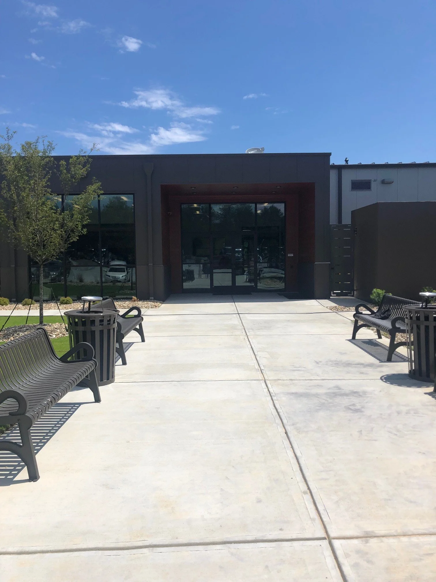 Outdoor seating area with benches and tables, trees, and a building with large glass doors under a clear blue sky.