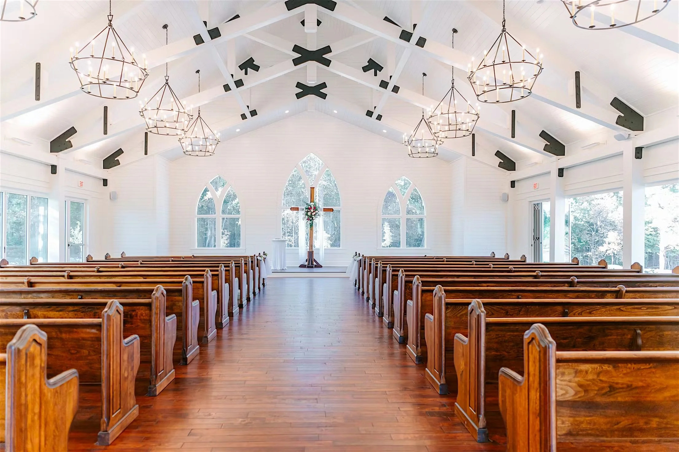 Interior of a bright church with wooden pews, large arched windows, a cross at the front, and chandeliers hanging from the ceiling.