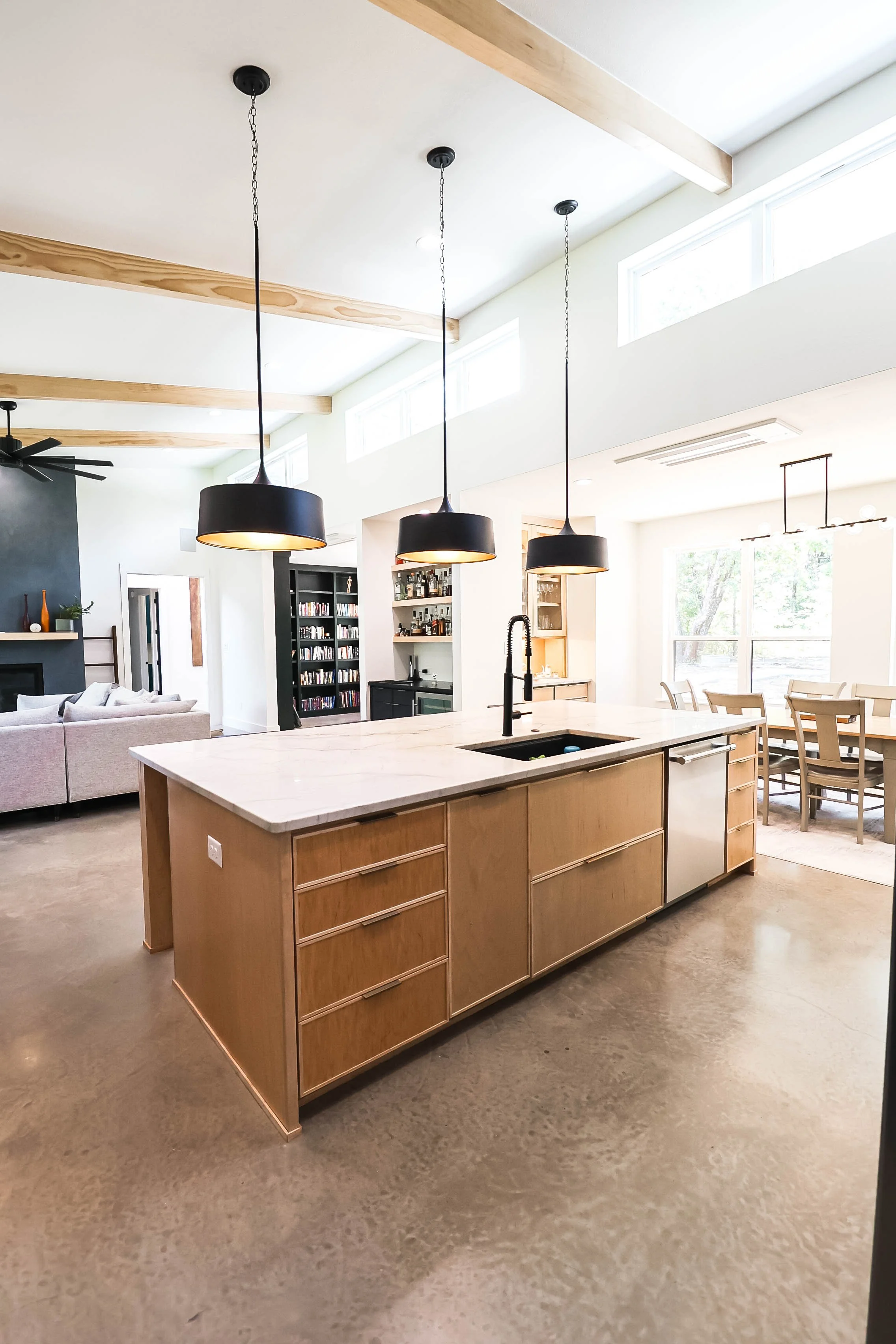 Modern open-plan kitchen and living room with light wood cabinets, black pendant lights, and large windows letting in natural light.