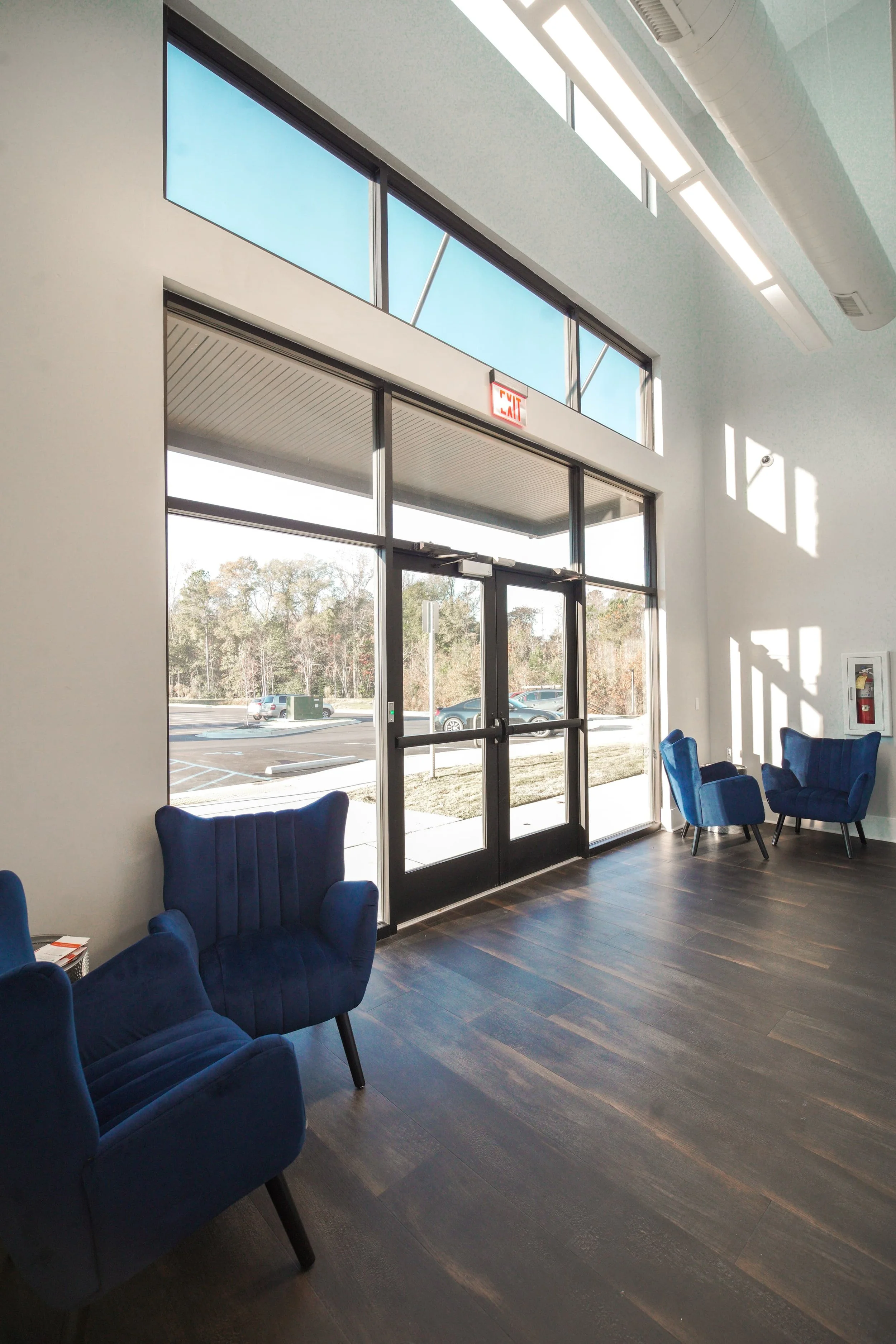 Interior of a modern waiting area with blue cushioned chairs, large glass windows, sunlight casting shadows, and a parking lot outside.