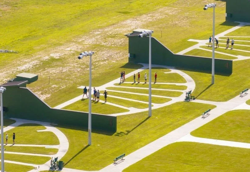Aerial view of a public park with lawn bowling grounds, paved walkways, benches, and people walking and socializing.