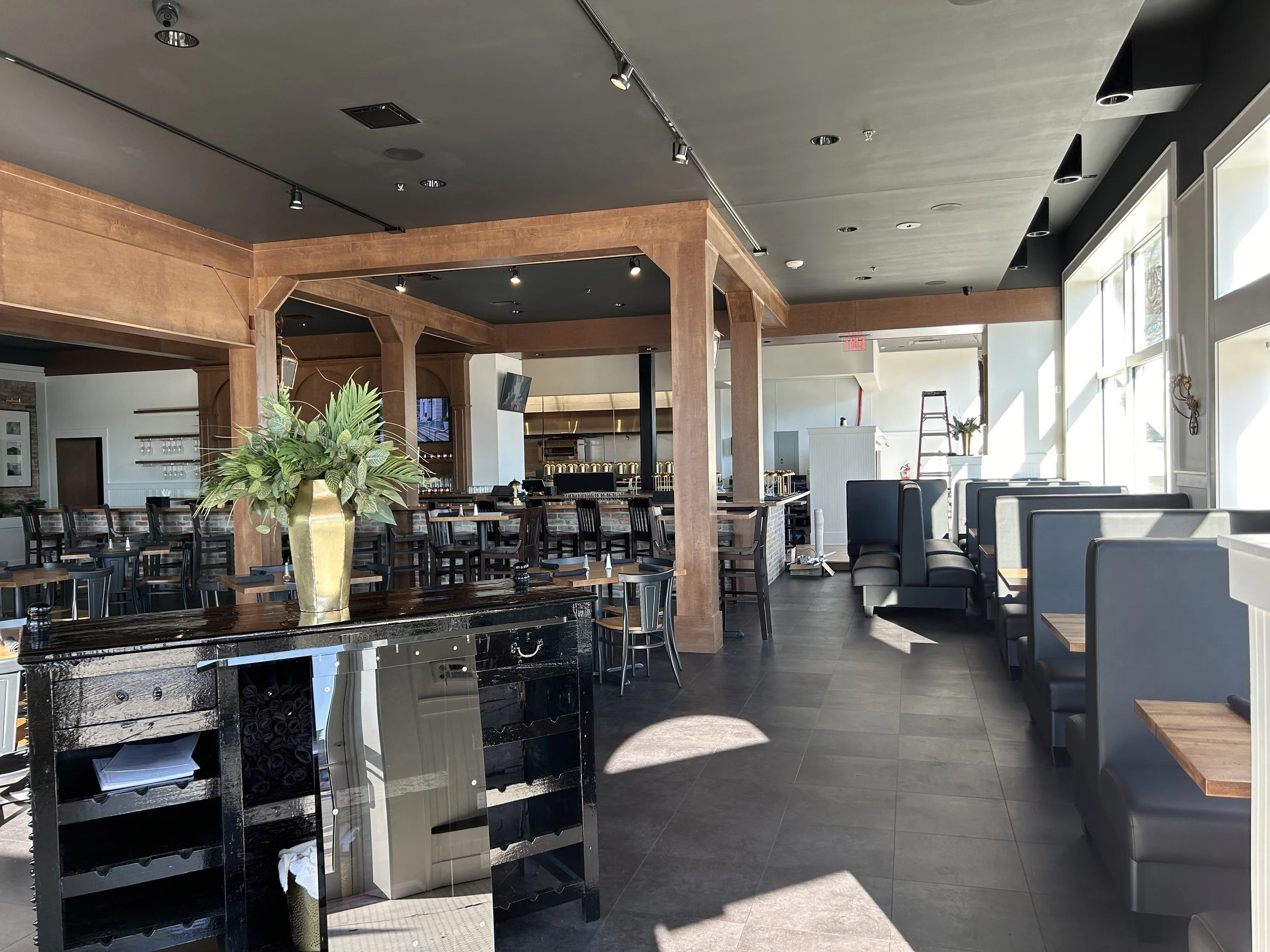 Interior of a restaurant or cafe with tables and chairs, a large plant in a gold vase, and a bar area in the background. The ceiling has track lighting and large windows allow natural light to fill the space.