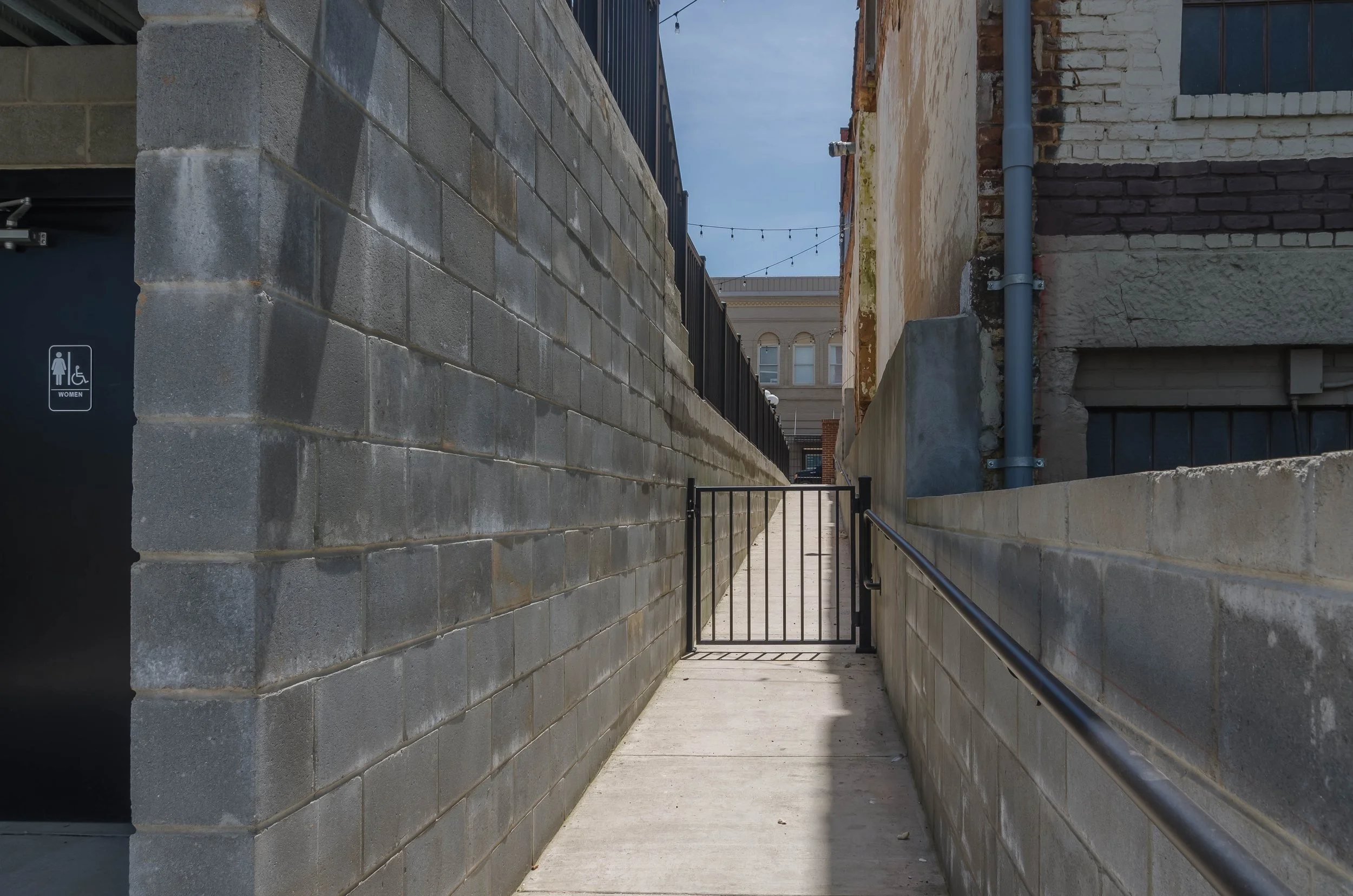 An alleyway with brick and concrete walls, a small gate at the end, a handrail on the right side, and string lights overhead under a partly cloudy sky.