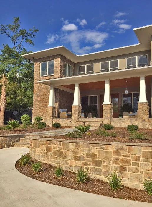 Front view of a modern two-story house with a stone exterior and large porch, surrounded by landscaped yard with small plants and a curved stone wall, under a blue sky with scattered clouds.