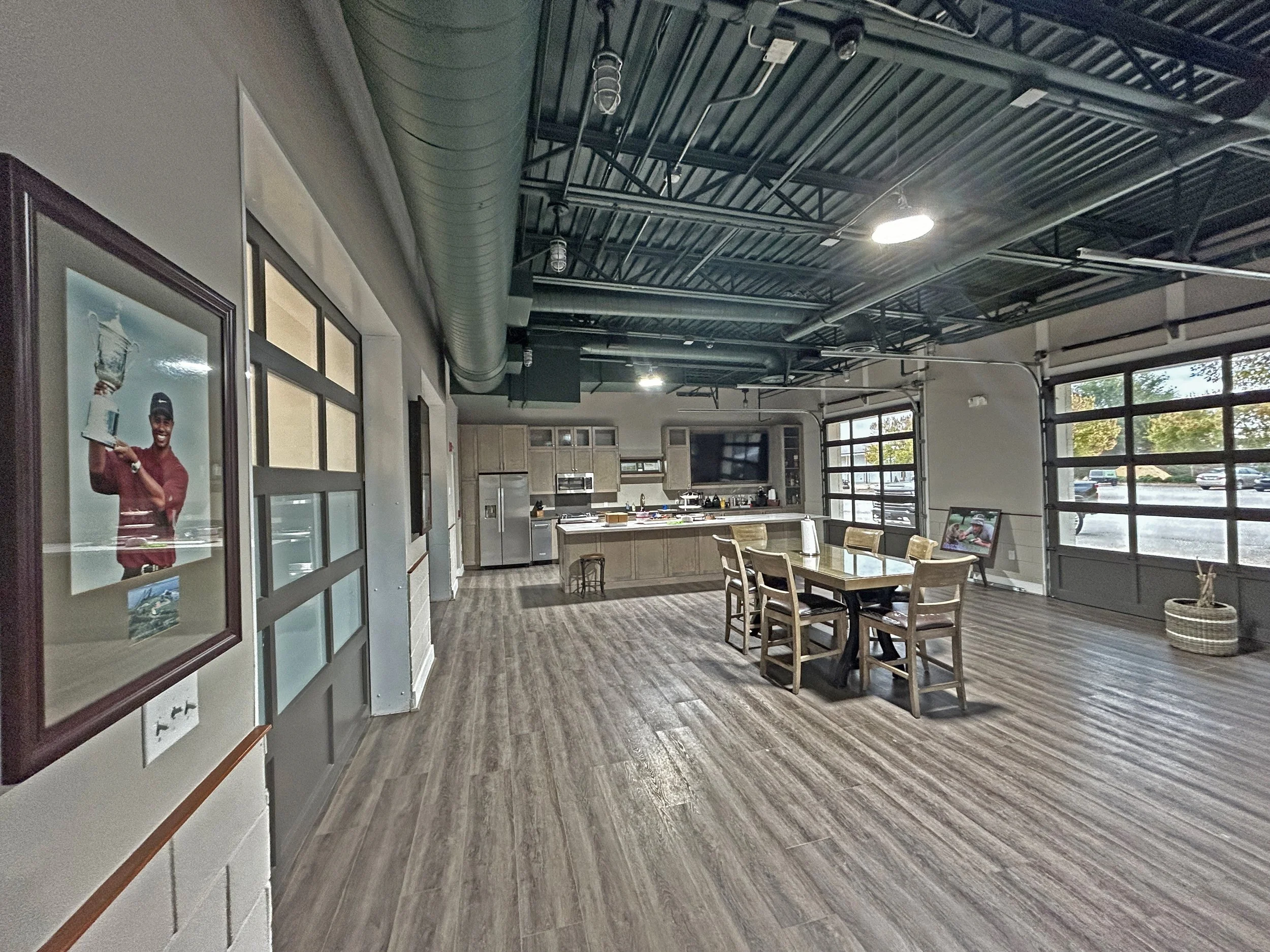 Interior of a modern industrial-style space with a high open ceiling featuring exposed ductwork and black metal framing, a large window allowing natural light, and a wood floor. There are tables and chairs in the foreground, a kitchen area in the background, and framed photographs on the wall.
